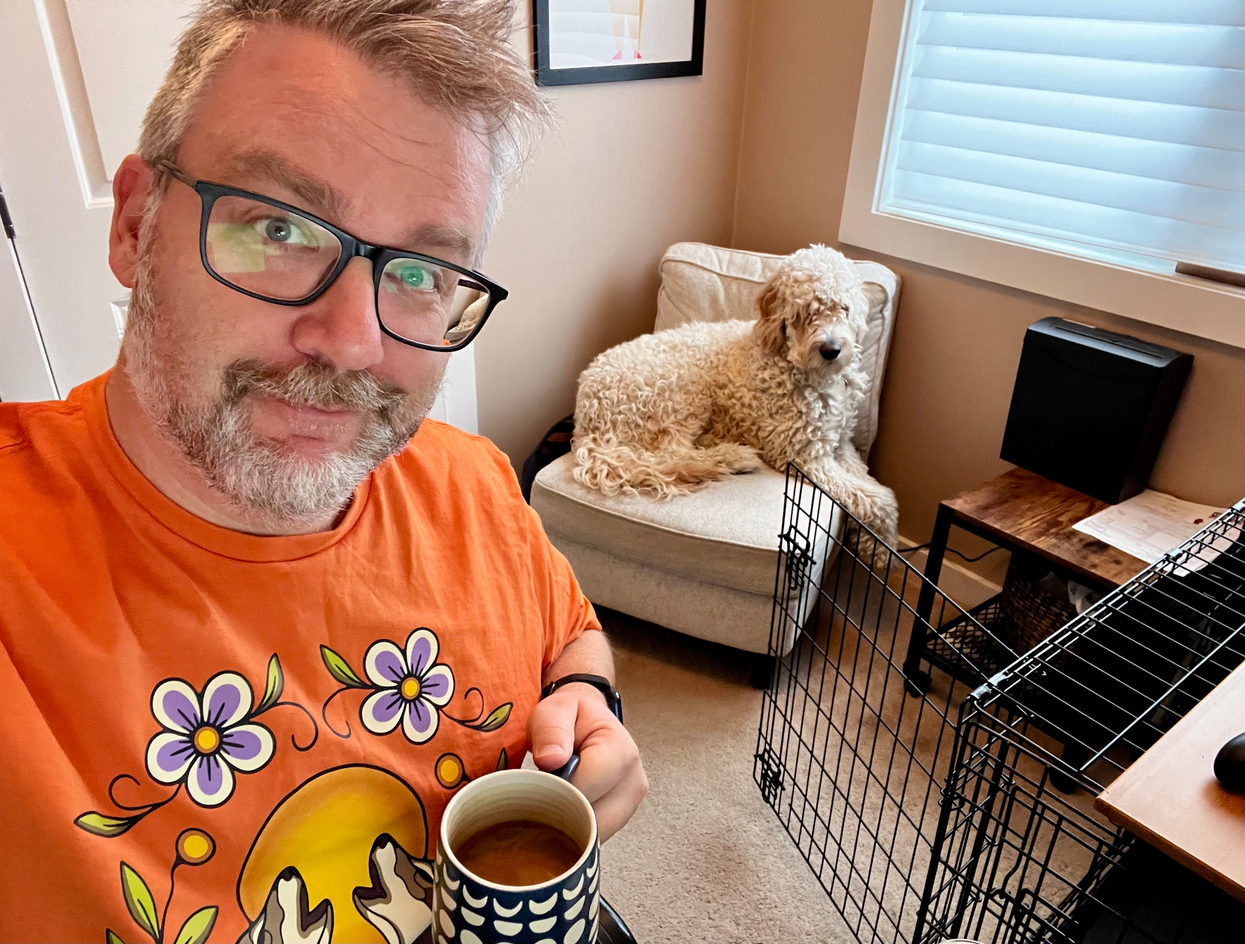 A person wearing an orange shirt with a floral design holds a cup of coffee while a fluffy dog sits on a chair in a room with a pet crate and a small table.