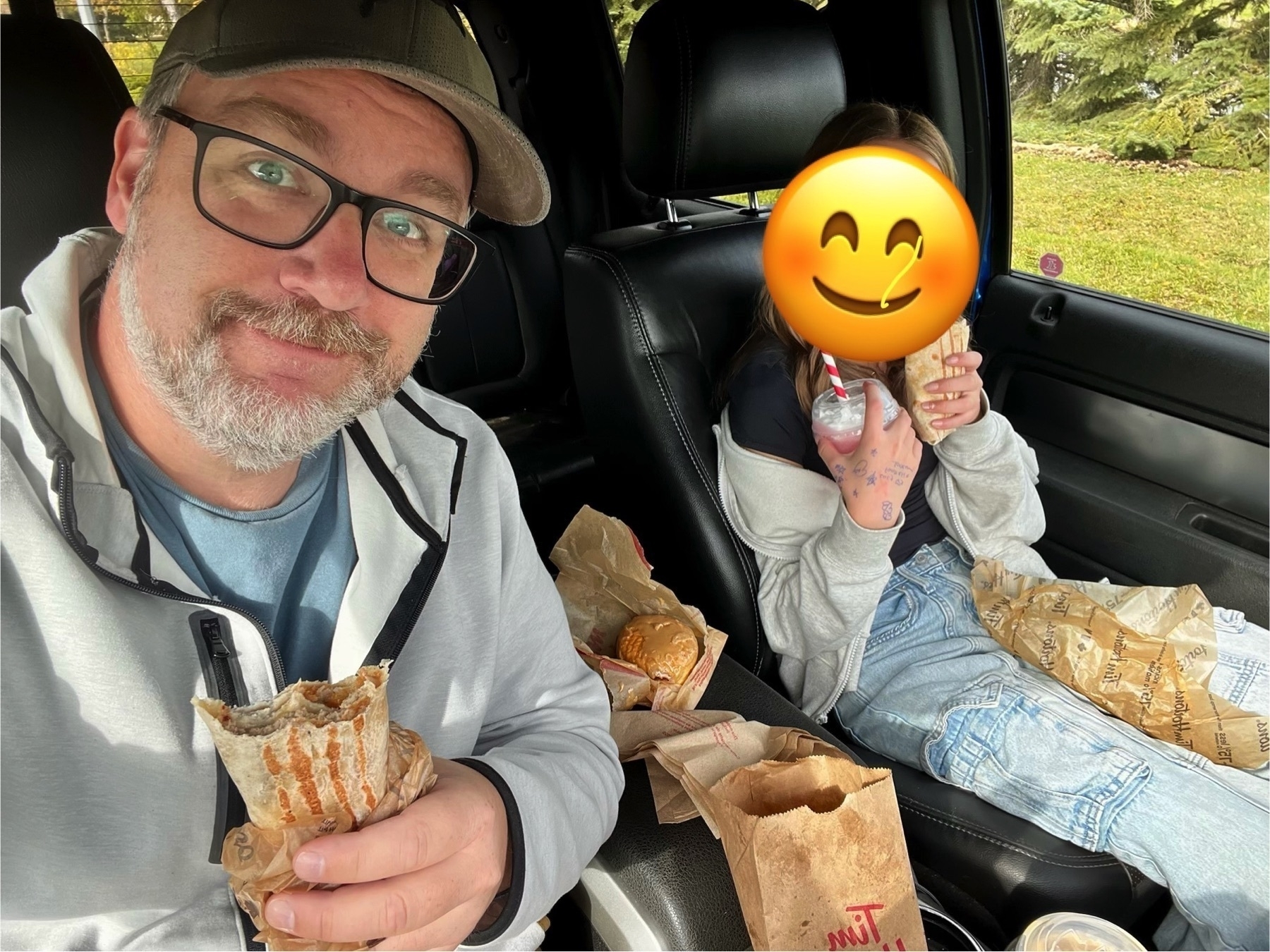 A person with glasses and a smiling emoji face over another person are enjoying fast food inside a car.