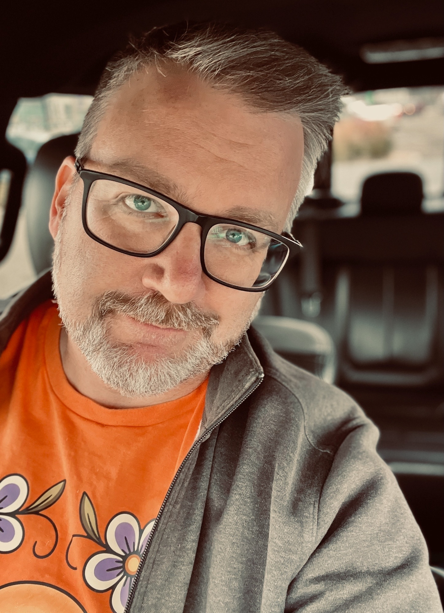 A man with glasses and a beard is sitting in a car, wearing a gray jacket over an orange shirt with floral designs.