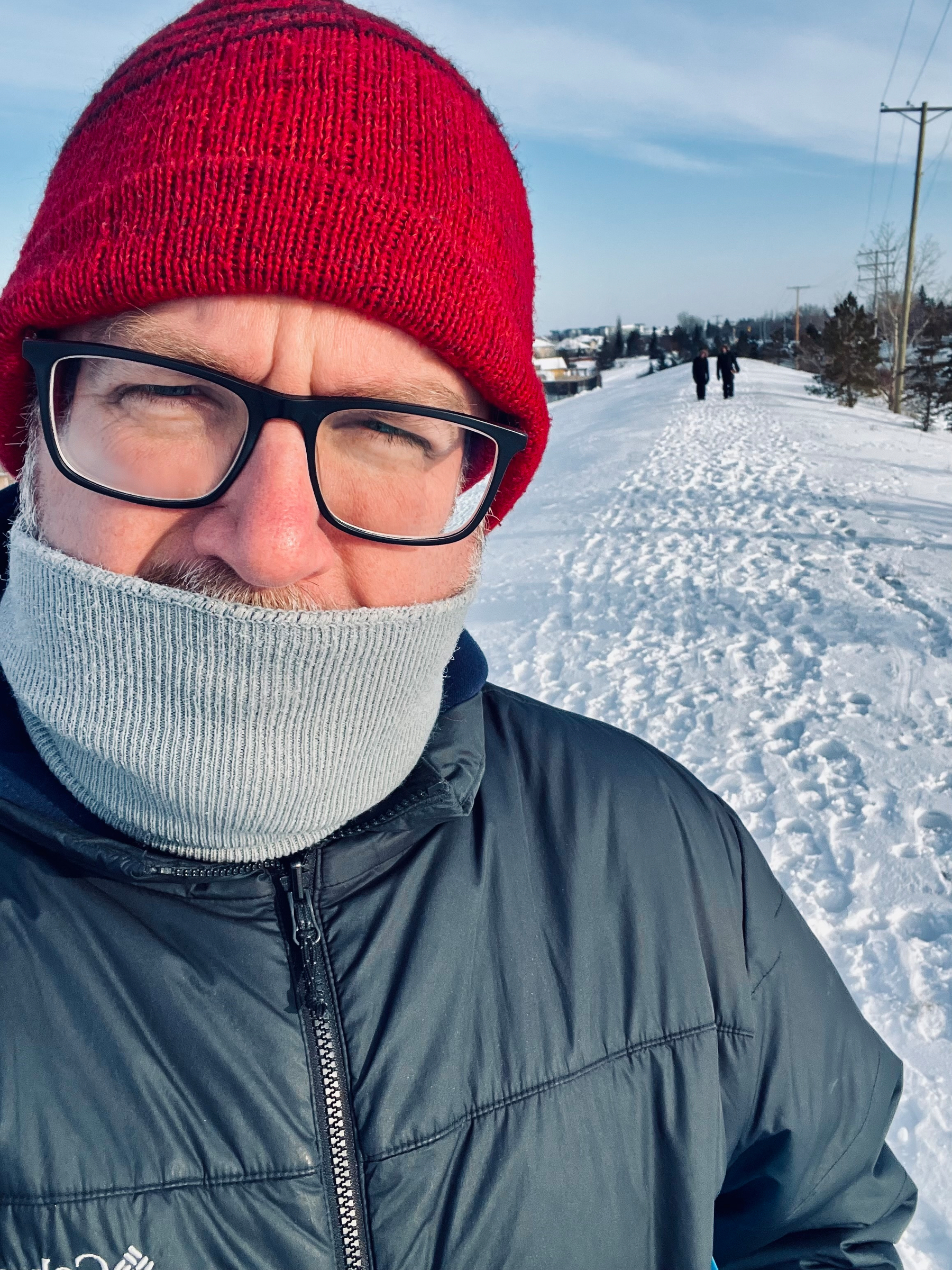 A person wearing a red beanie, glasses, and a scarf around their face is standing on a snow-covered path.