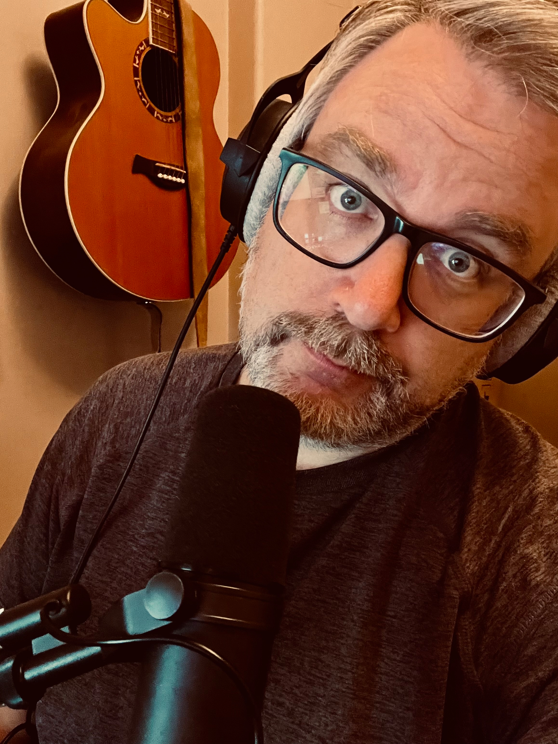 A person wearing headphones and glasses is speaking into a microphone, with a guitar hanging on the wall behind them.