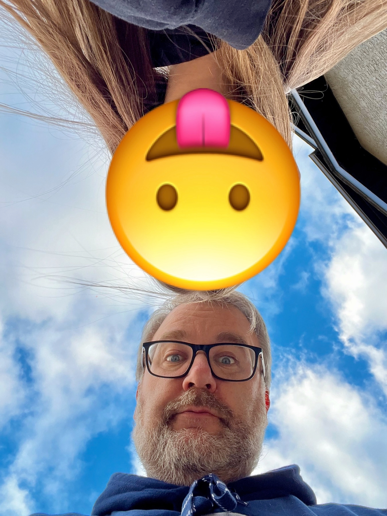 A person with glasses is looking down at the camera with a bright sky in the background, and a face with a playful tongue-out emoji covers their face.