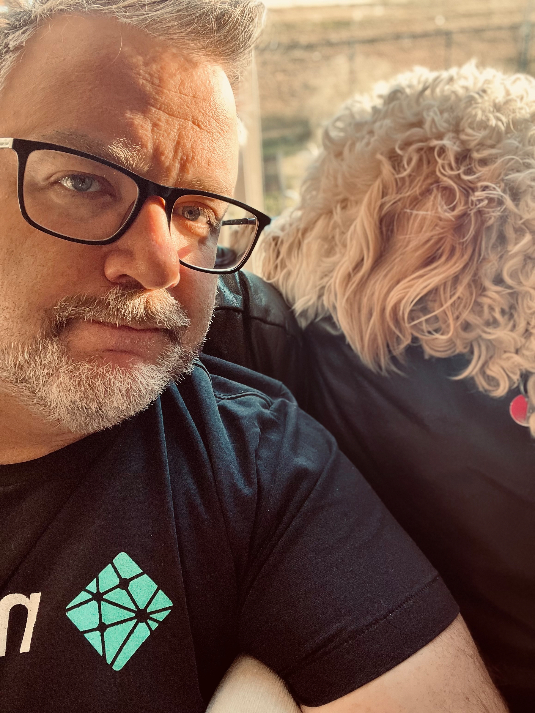 A man wearing glasses and a black t-shirt is sitting next to a curly-haired dog.
