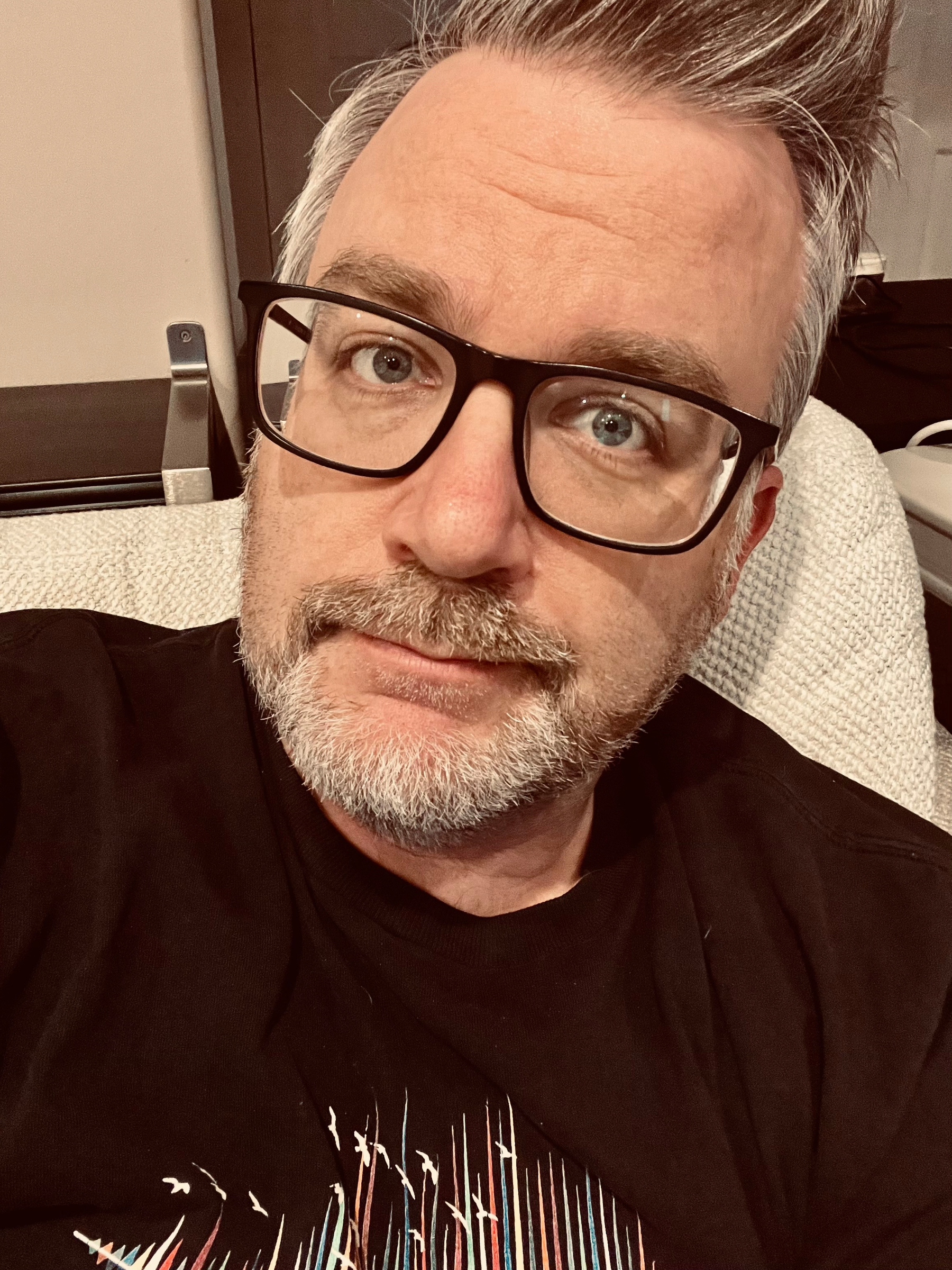 A person with glasses and a beard is reclining on a textured surface, looking at the camera.