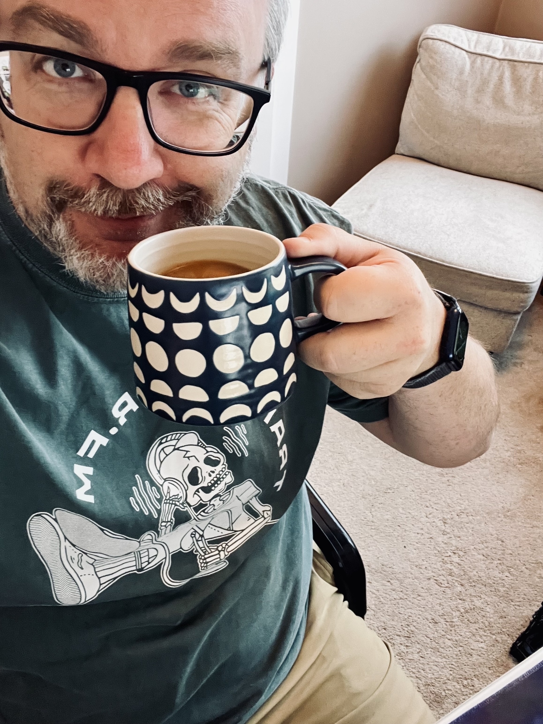 A person wearing glasses holds a patterned mug of coffee, smiling, while seated in a cozy environment.