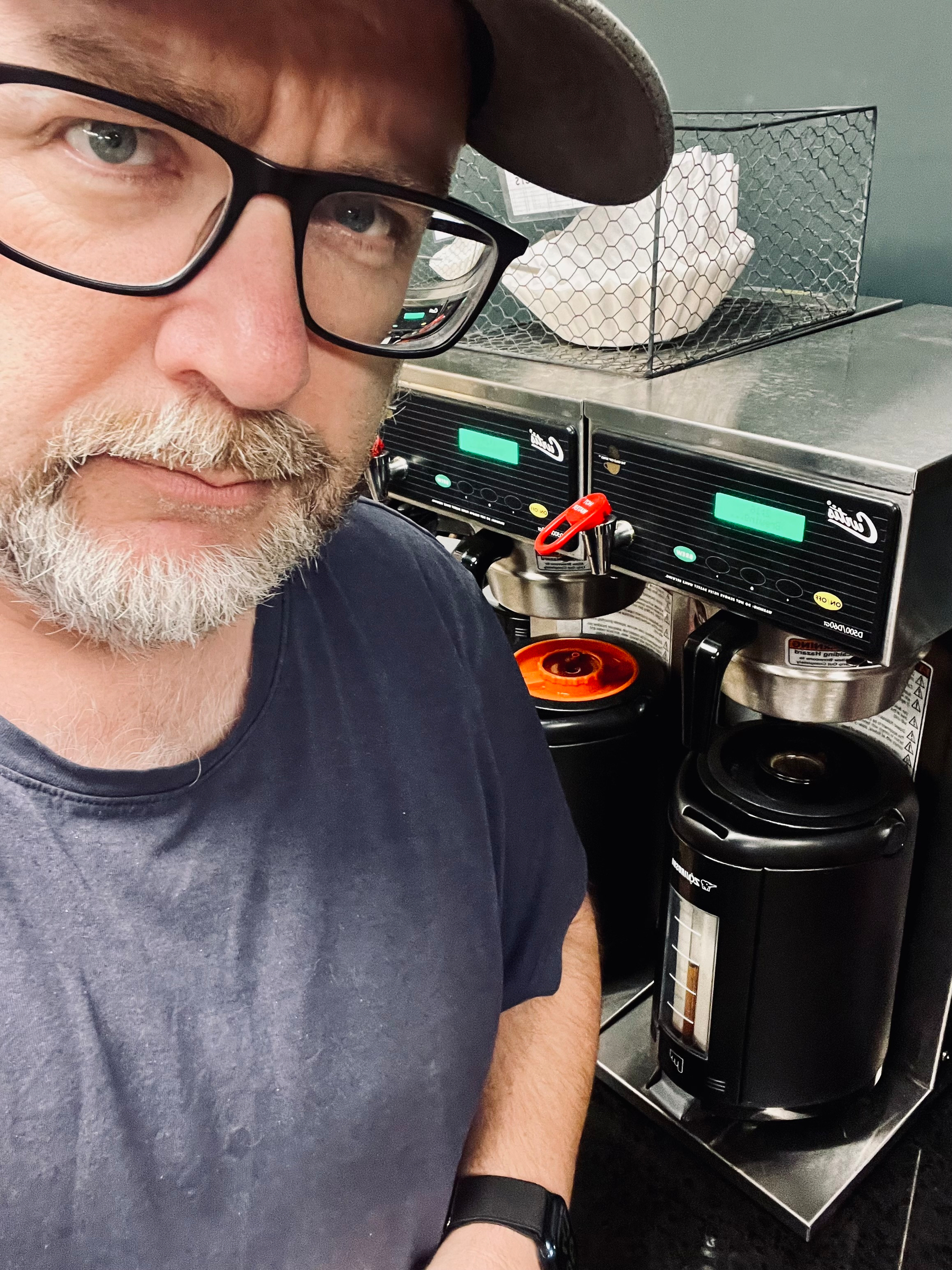 A person wearing glasses and a cap appears concerned in front of a commercial coffee machine with multiple pots.