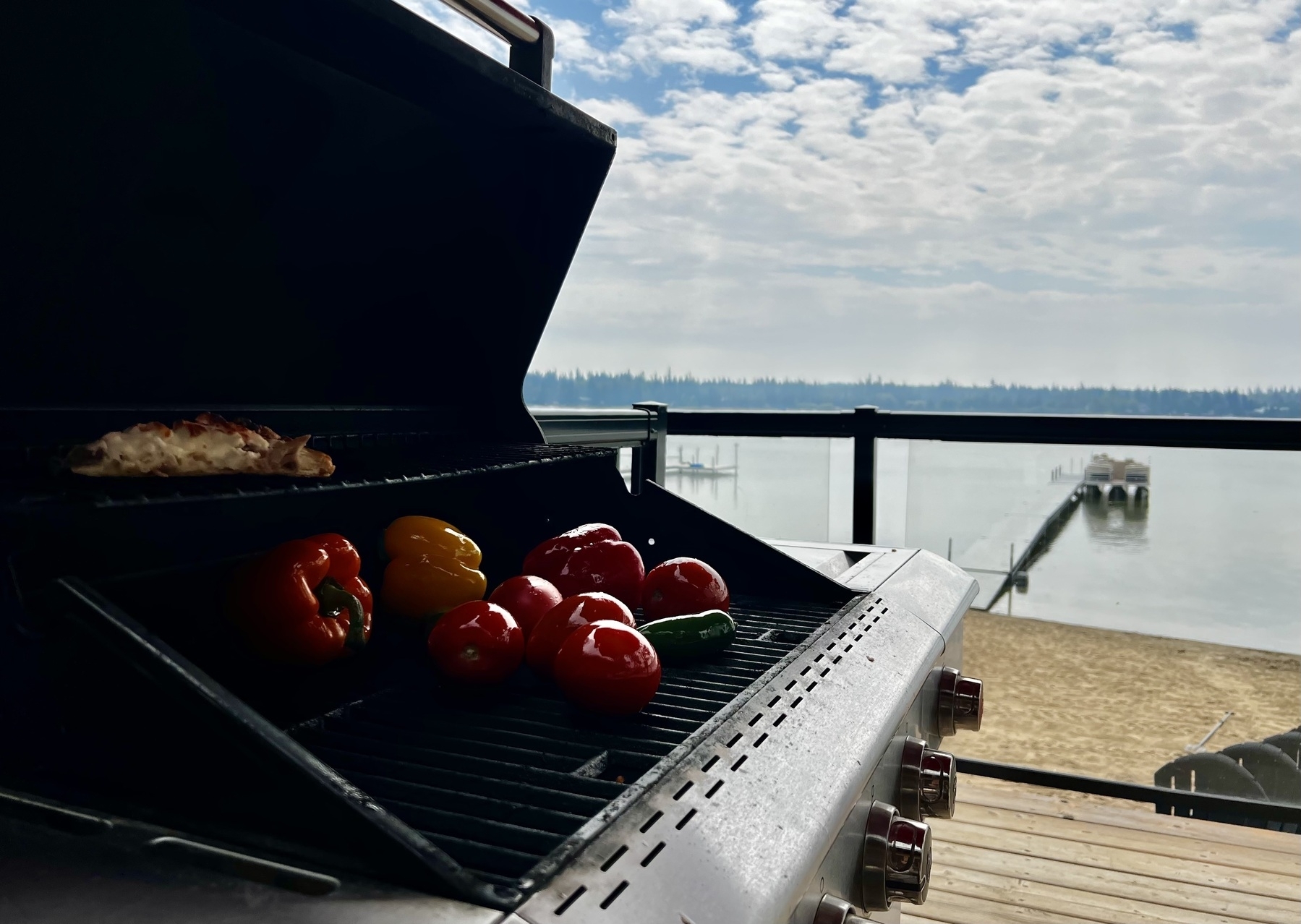 A bbq in the foreground has peppers, tomatoes, onions, and more on it cooking. A lake is in the background. 