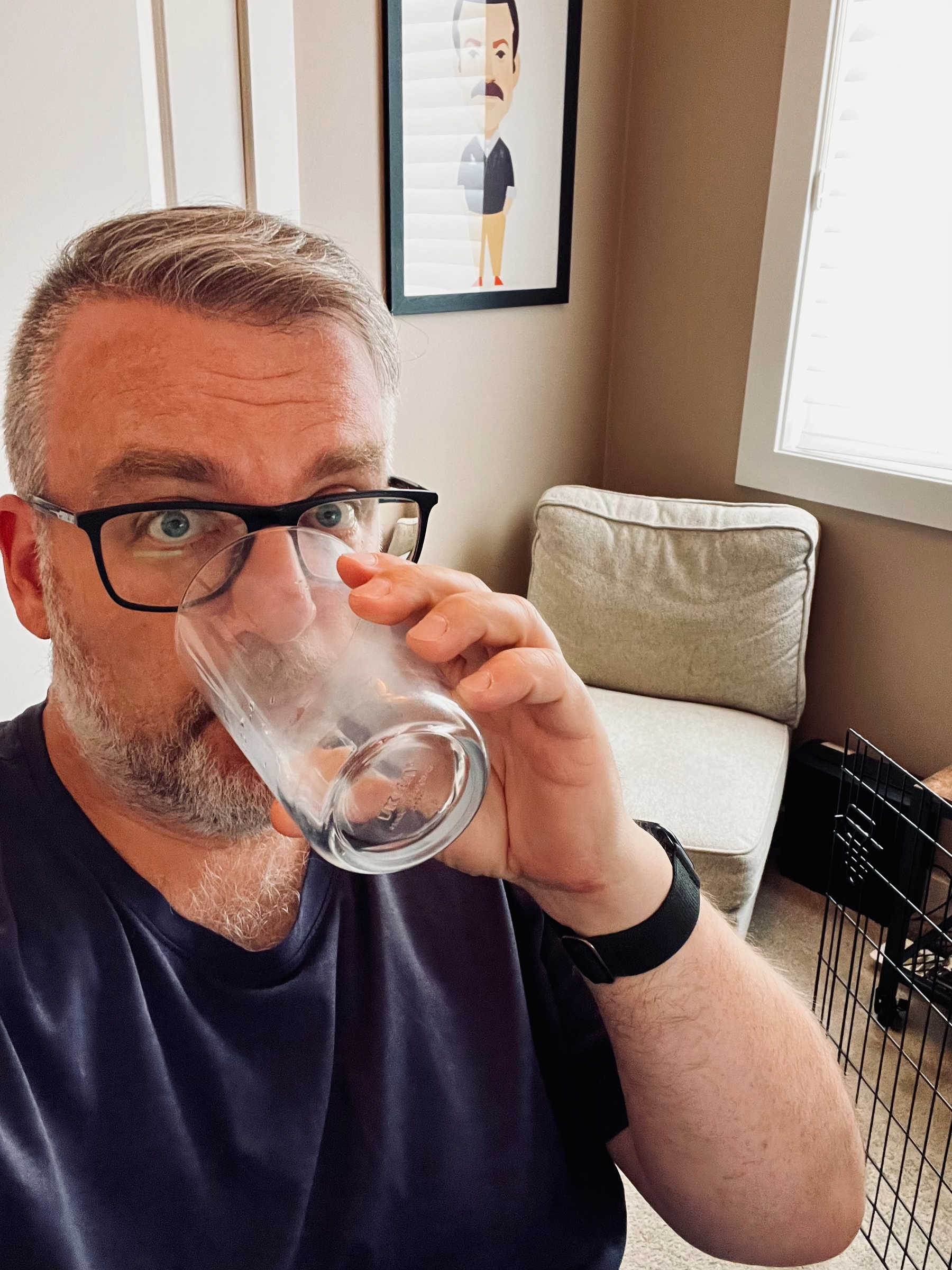 A man with glasses is drinking from a clear glass while sitting in a room with a framed cartoon portrait of Ted Lasso on the wall and a beige chair in the background.