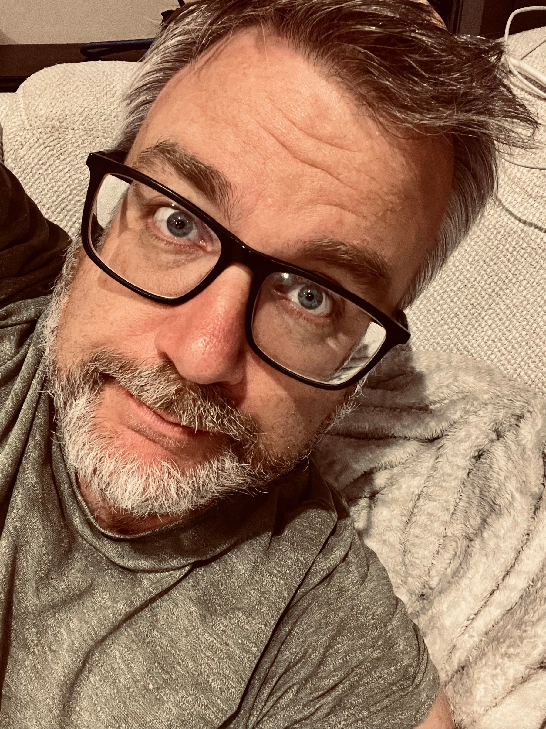 A middle-aged man with gray hair, glasses, and a beard is lying on a light-colored couch, looking directly at the camera.