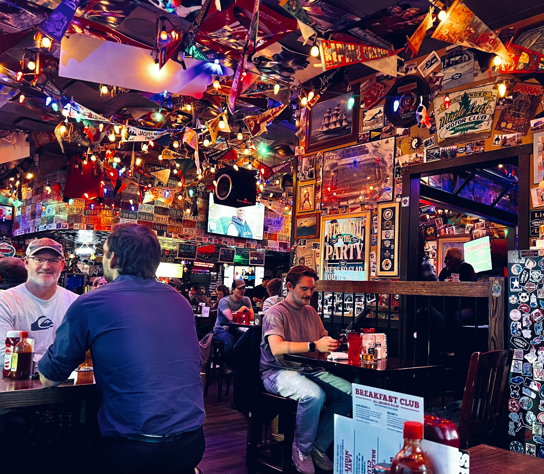 A lively, colorful bar or restaurant interior filled with people dining and decorated with numerous flags, posters, and string lights.