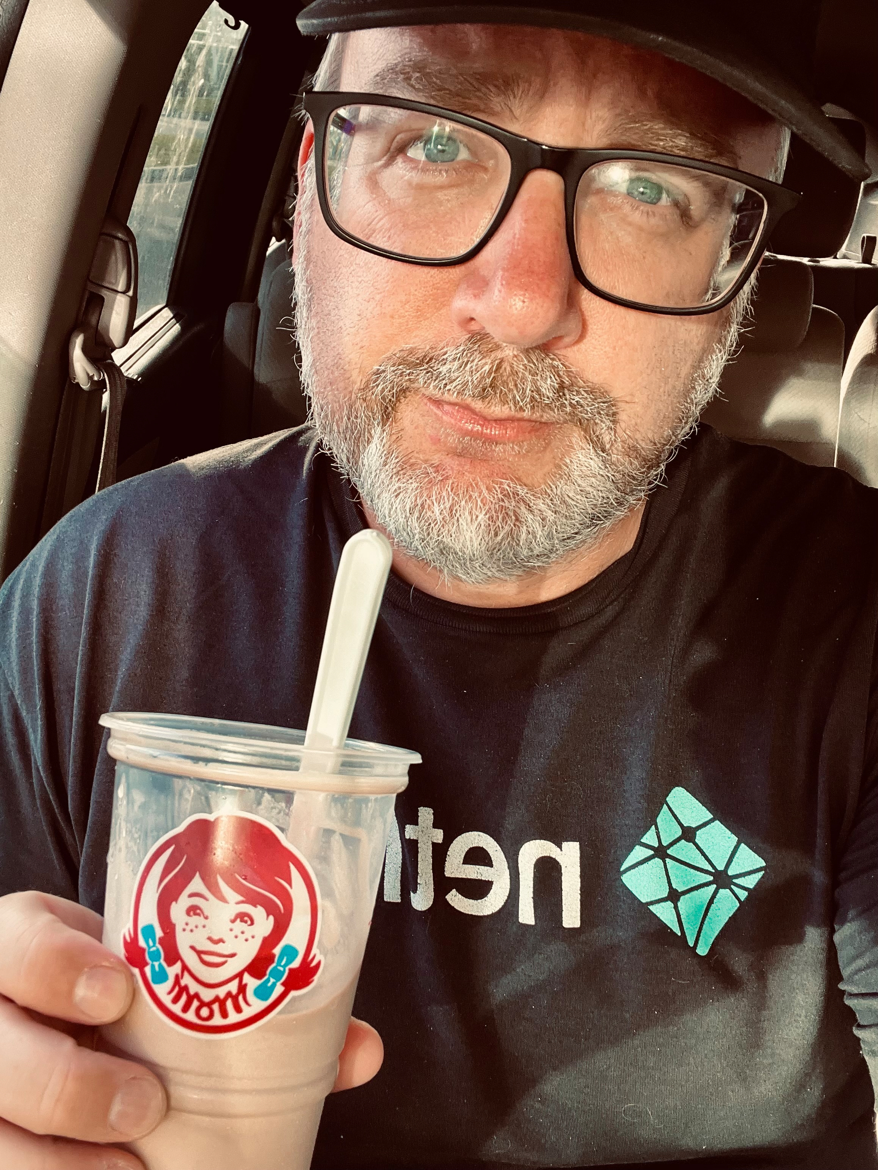 A bearded man wearing glasses and a cap is sitting in a car, holding a plastic cup with a straw that features a logo of a girl with red braids.