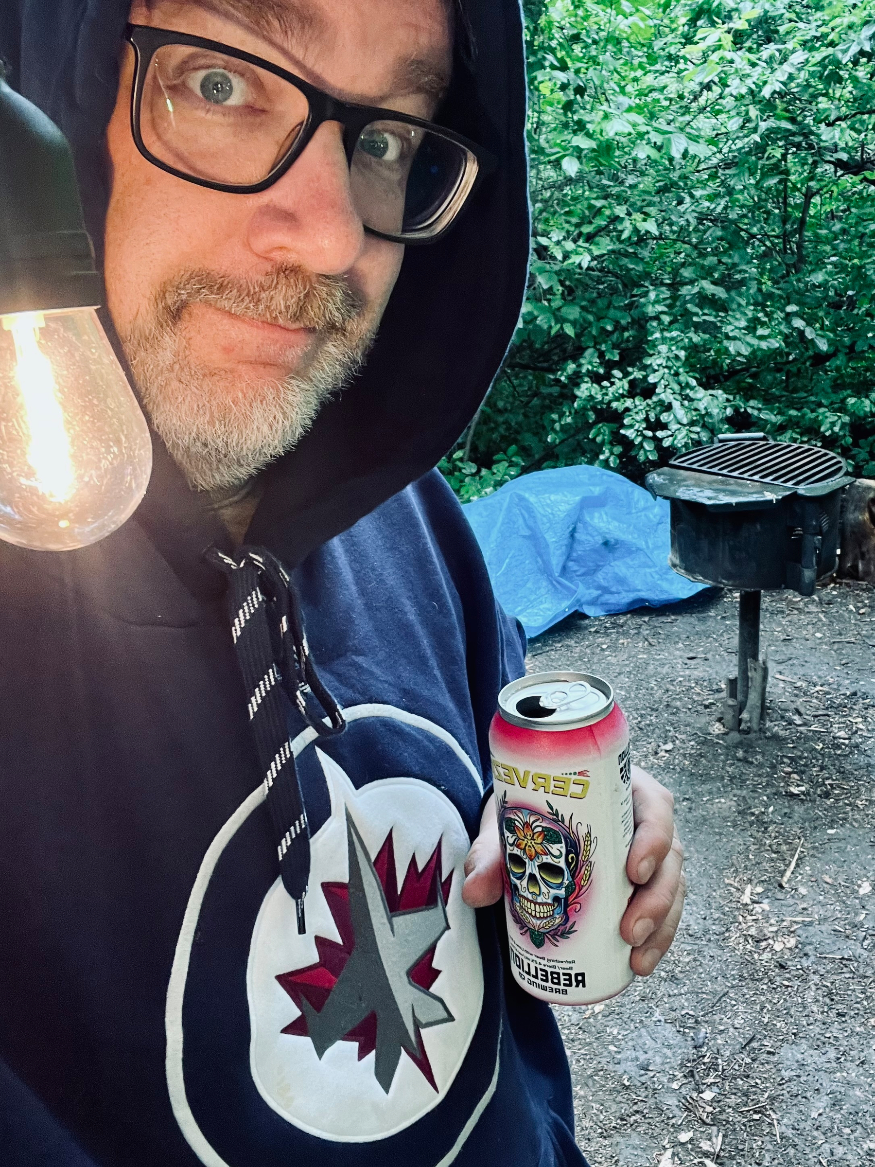 A man wearing glasses and a hoodie with a logo is holding a can of beverage near a lit bulb and an outdoor grill in a wooded area.