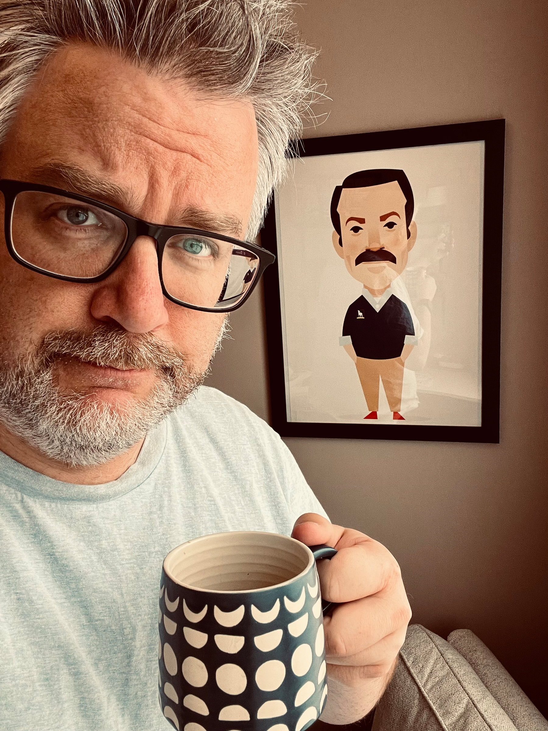 A man with glasses holds a patterned mug in front of a wall with a framed cartoon portrait of Ted Lasso in the background.
