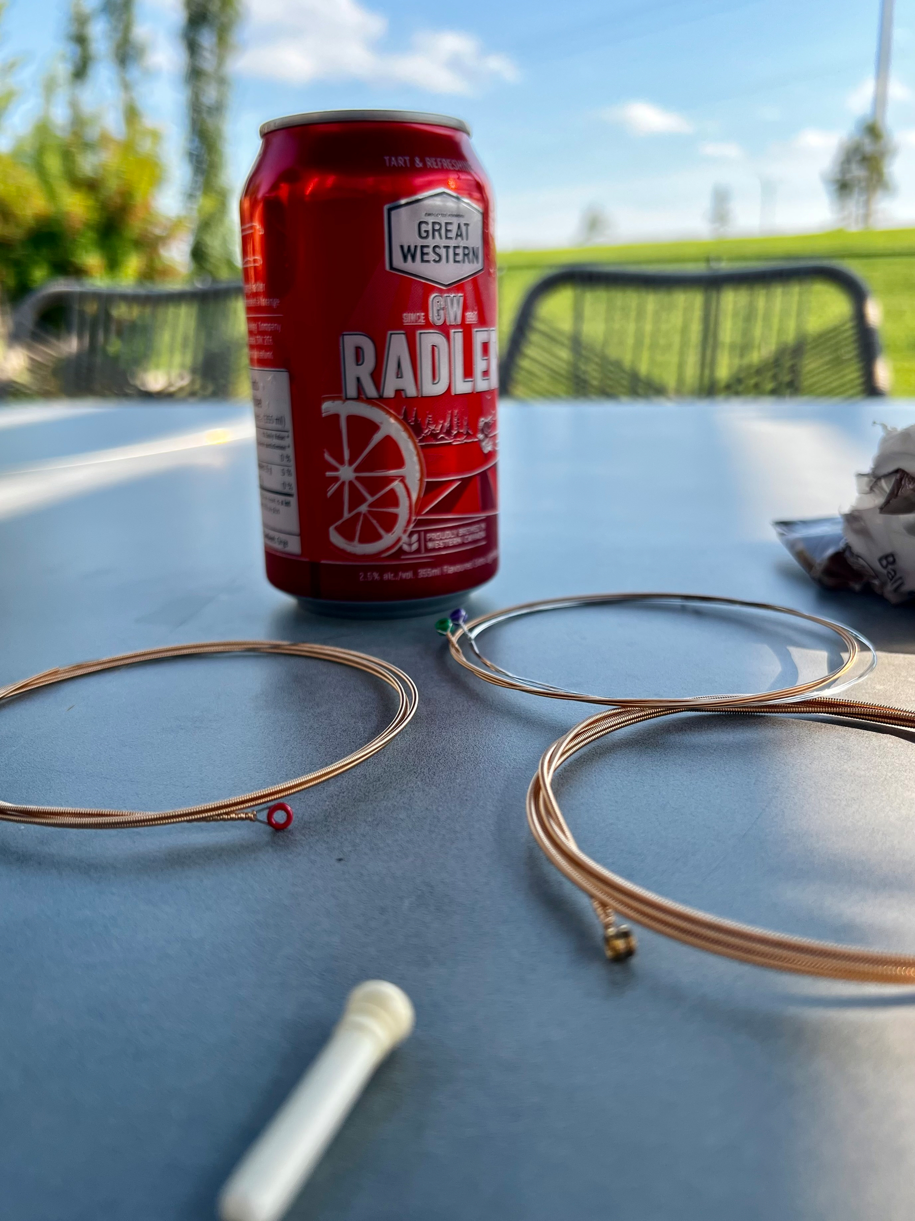 A can of Great Western Radler sits on a table alongside three coiled guitar strings and a white guitar bridge pin, with an outdoor setting visible in the background.