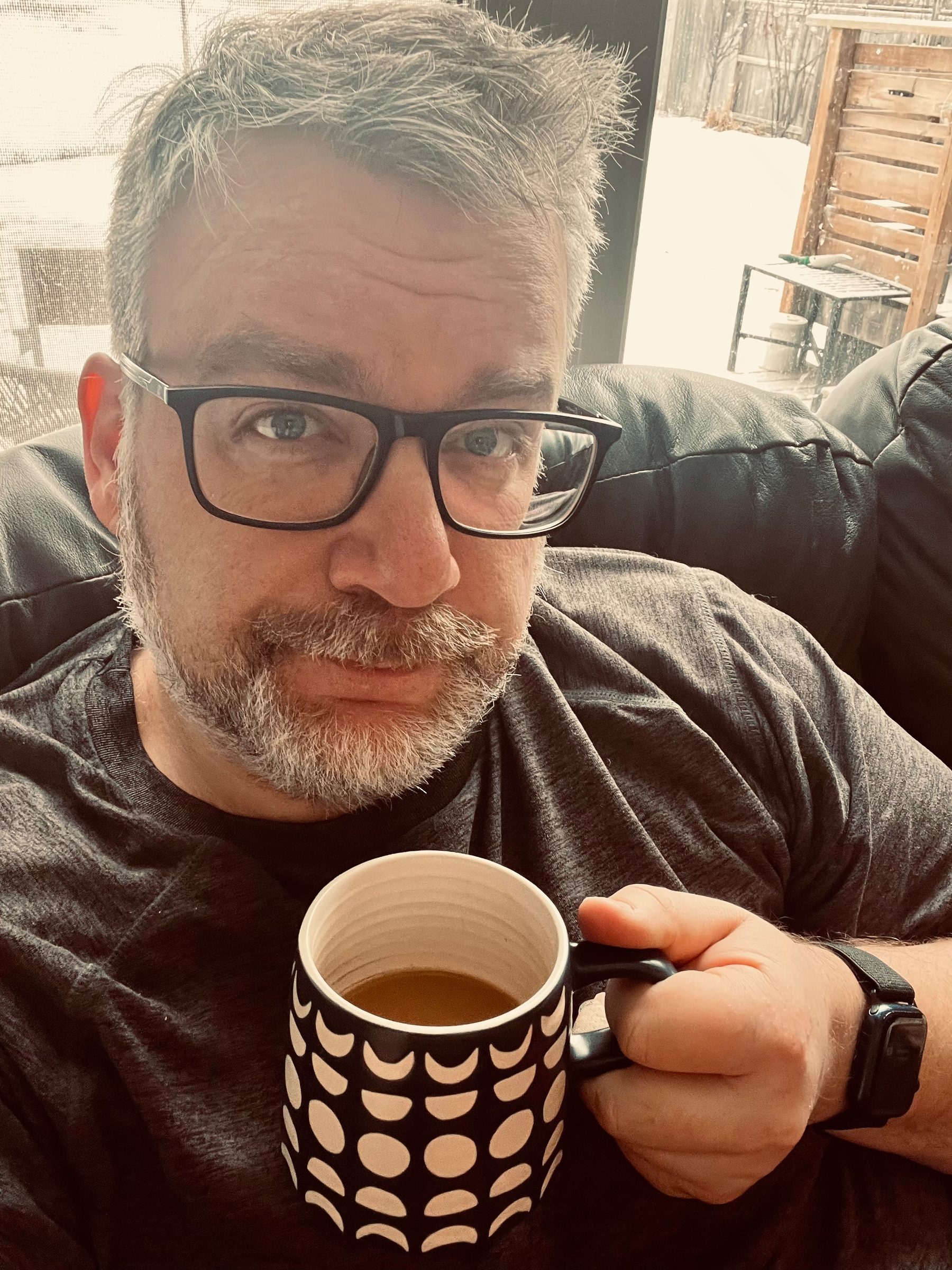 A person with glasses is sitting on a couch, holding a mug with a geometric black and white pattern.