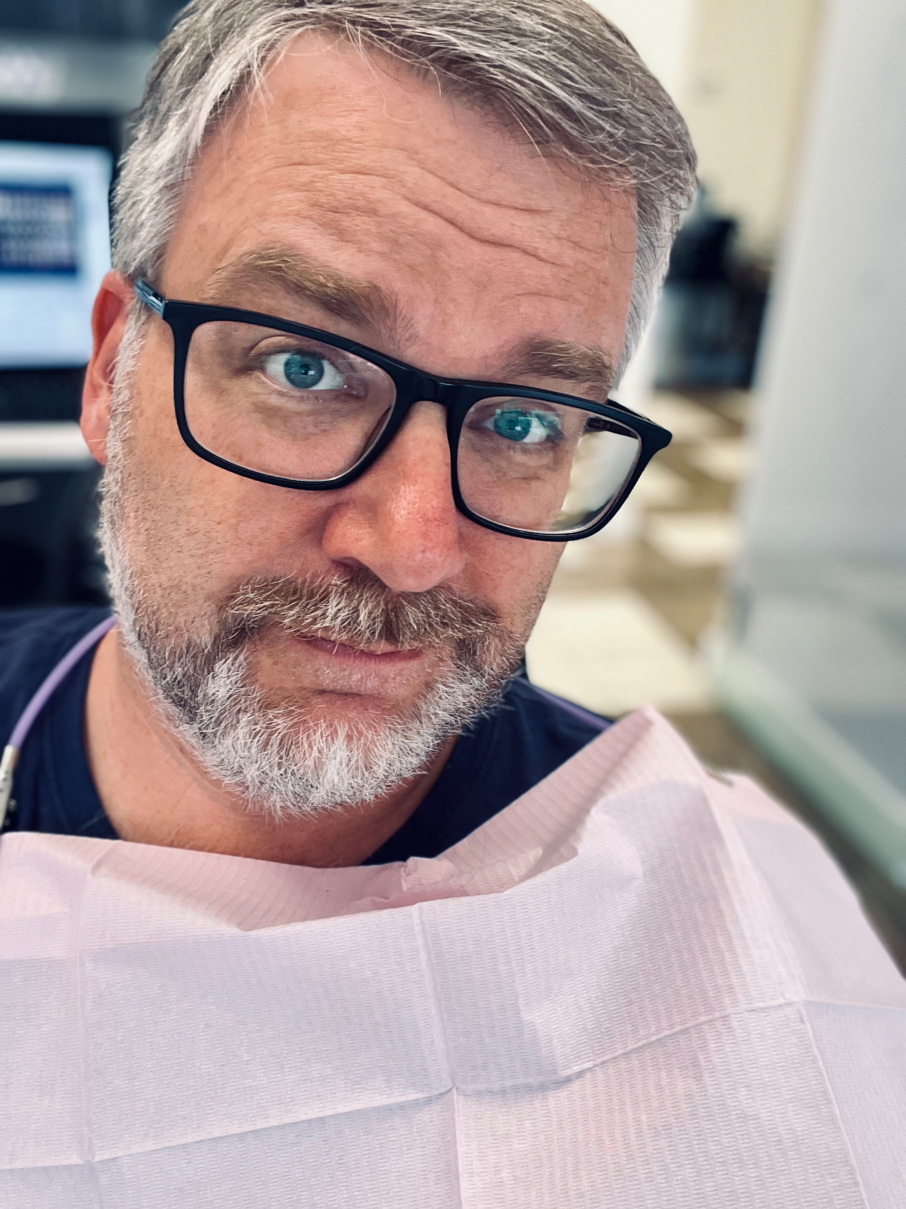 A man wearing glasses is sitting indoors with a draped cloth around his neck, perhaps at a dental clinic or similar setting.