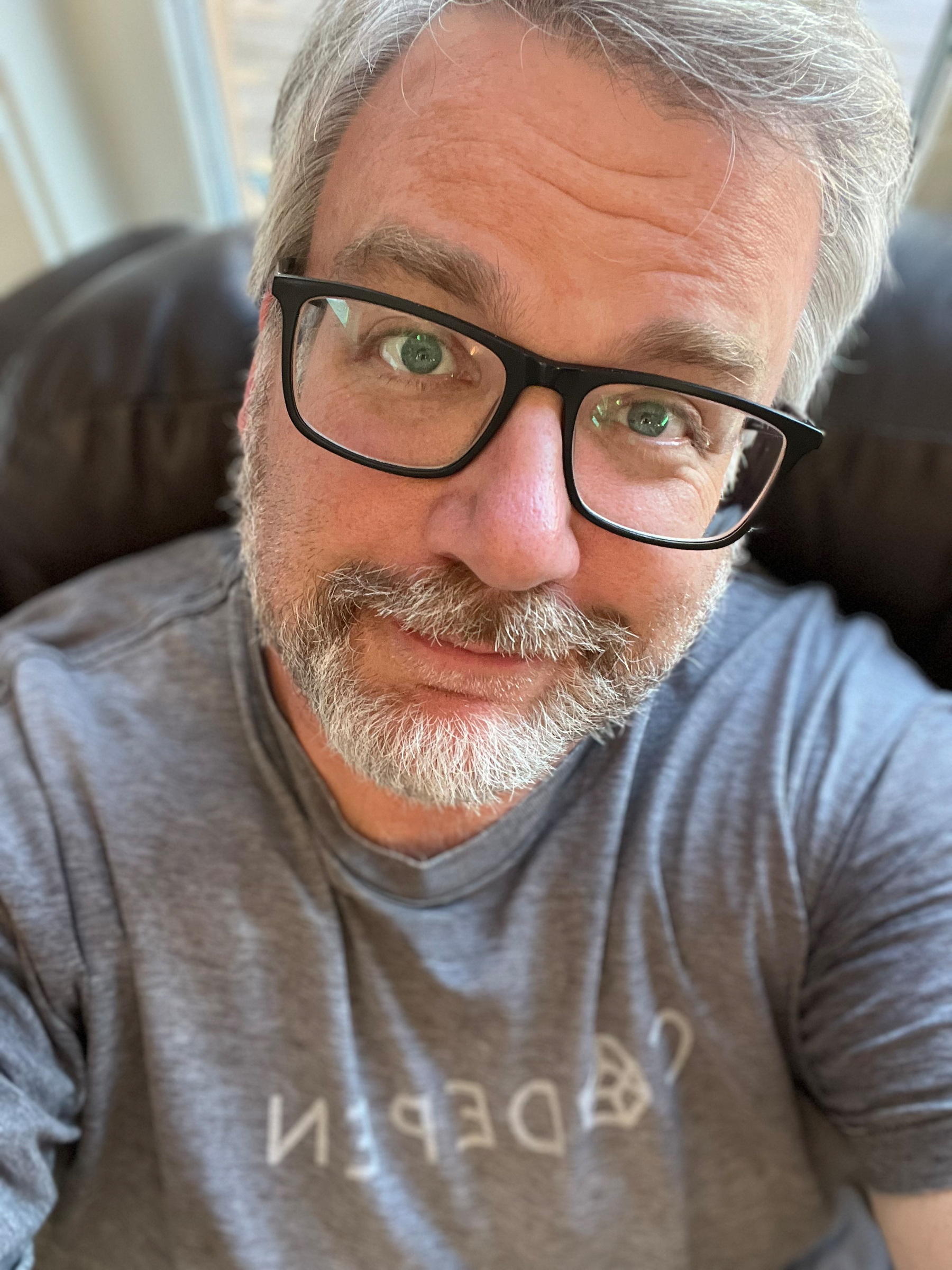 A person with glasses and a beard is wearing a gray shirt and looking at the camera.