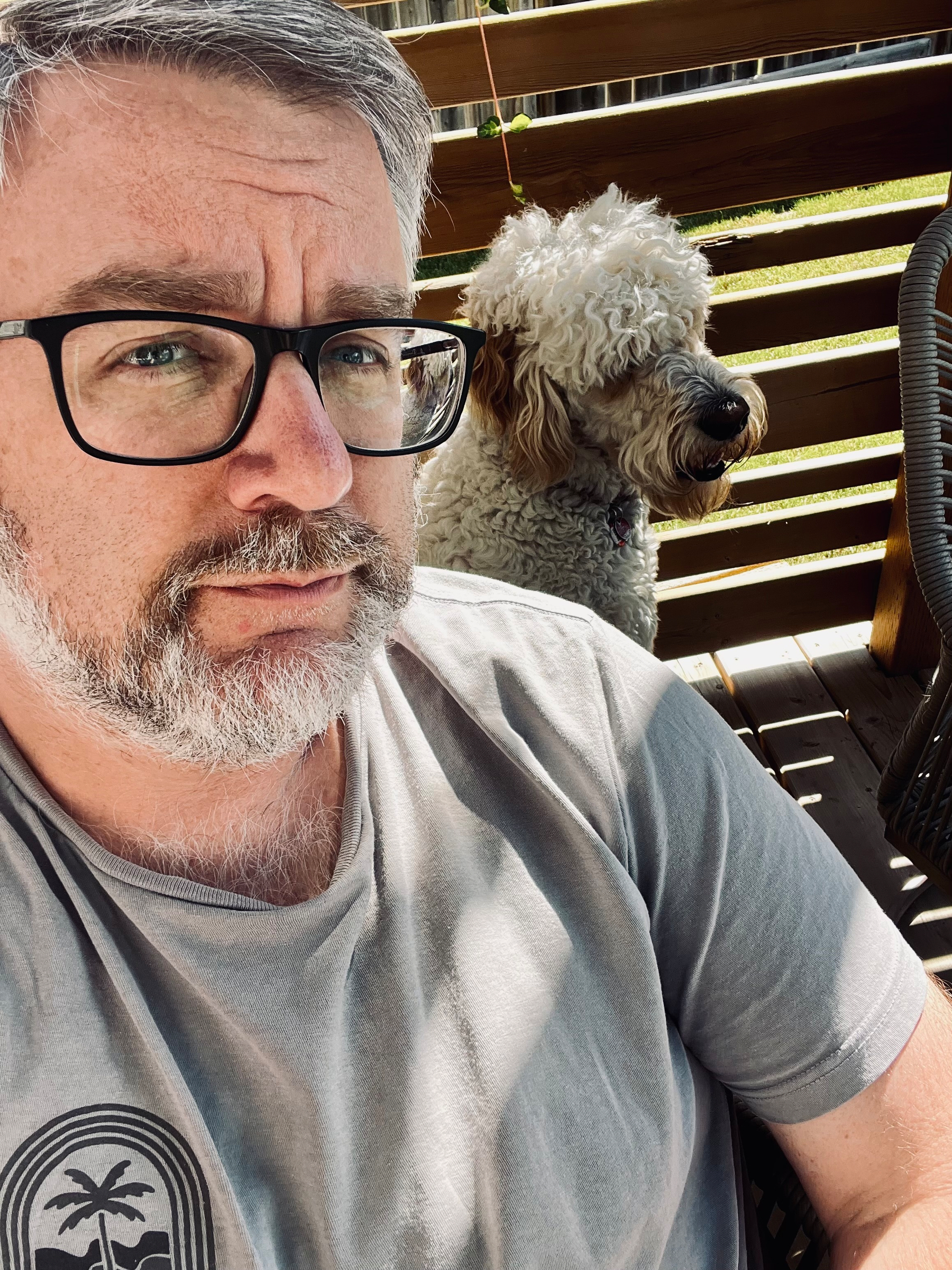 A man with glasses and a beard is sitting outside on a wooden deck with a curly-haired dog behind him.