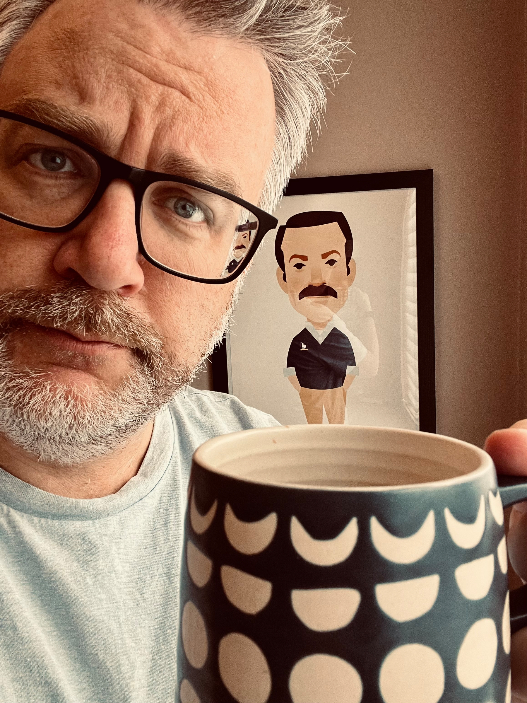 A person with glasses holds a patterned mug in front of a portrait of Ted Lasso in the background.