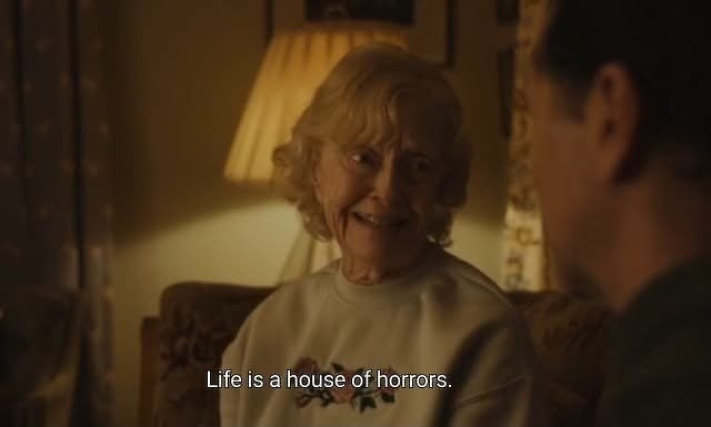 An elderly woman with curly blonde hair is sitting on a couch and speaking to a man, with the subtitle Life is a house of horrors. displayed at the bottom. From Widow’s Bay.