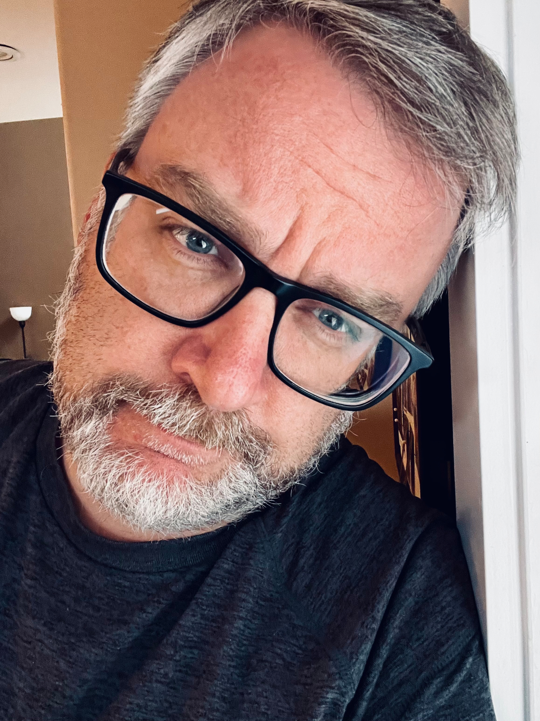 A bearded man with glasses leans against a wall, looking into the camera.
