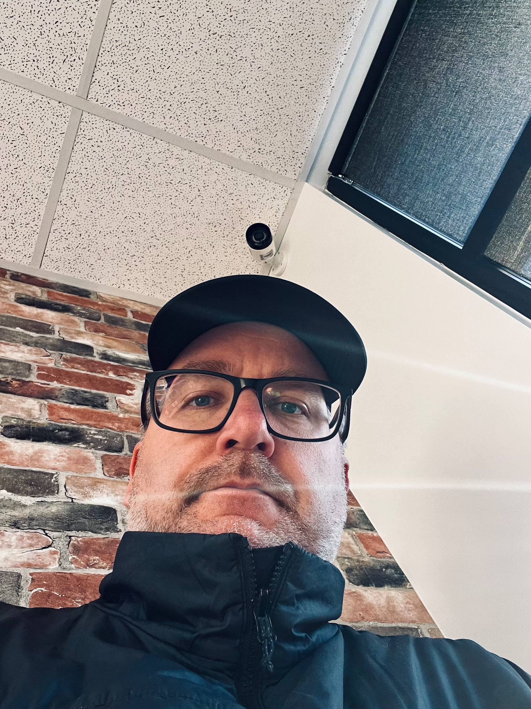 A person wearing glasses and a cap is looking at the camera from a low angle, with a ceiling and brick wall in the background.