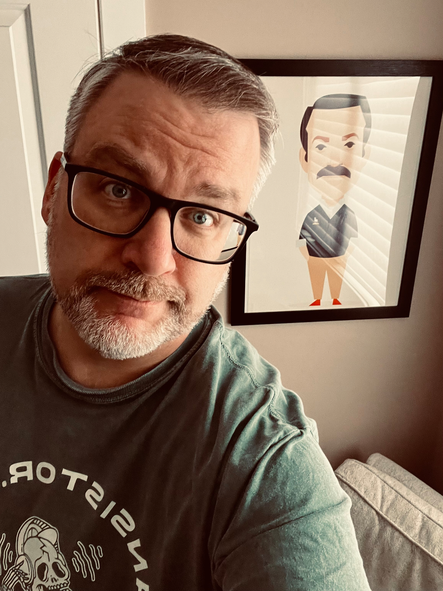 A man with glasses is looking at the camera while standing next to a framed cartoonish illustration of Ted Lasso. 