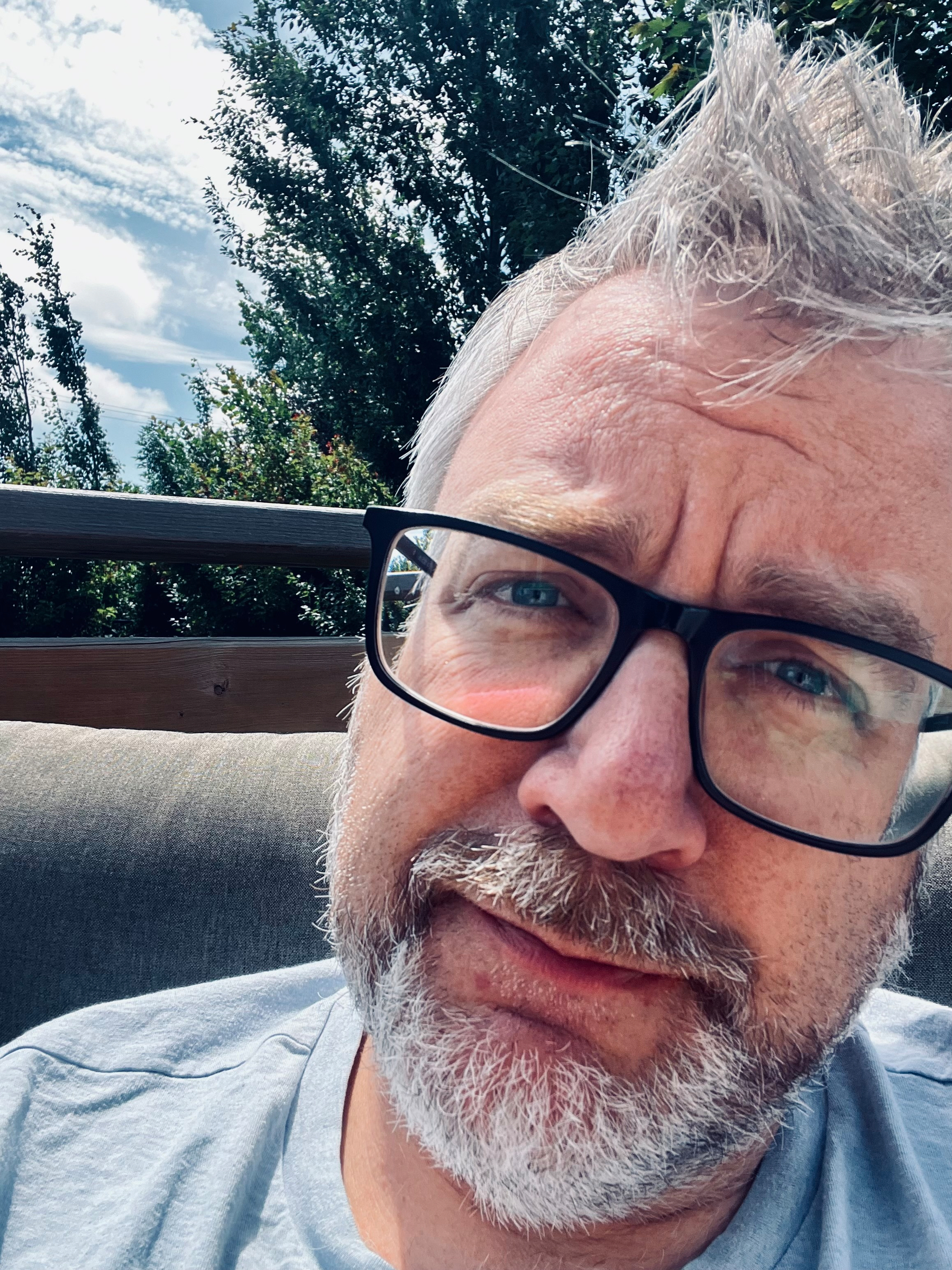 A man with gray hair, beard, and glasses is sitting outdoors on a couch or seat with trees and a partly cloudy sky visible in the background.