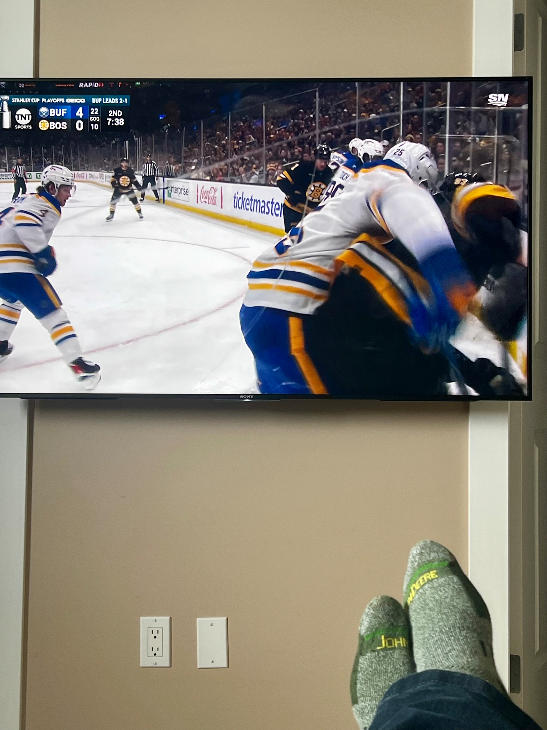 A person is watching a hockey game on TV while relaxing with their feet up, showing a match between the Buffalo Sabres and Boston Bruins.