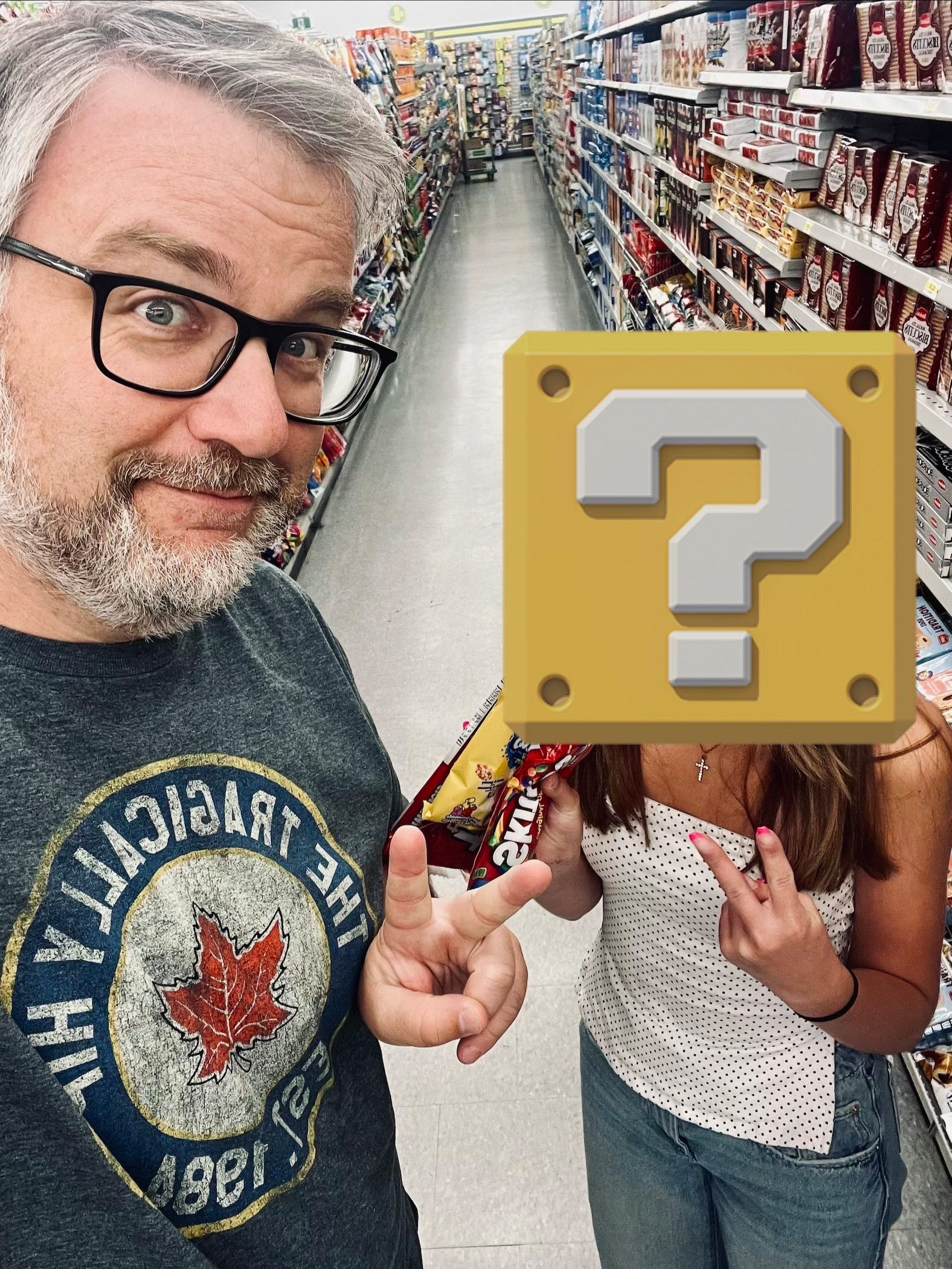 A man wearing glasses and a graphic t-shirt poses with his daughter in a grocery store aisle, both making peace signs.