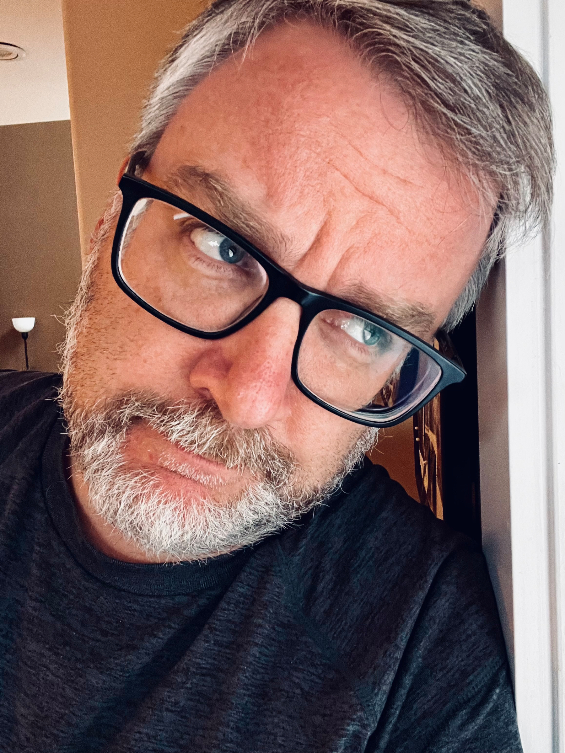 A man with glasses and a beard leans against a wall while looking to the side with a contemplative expression.