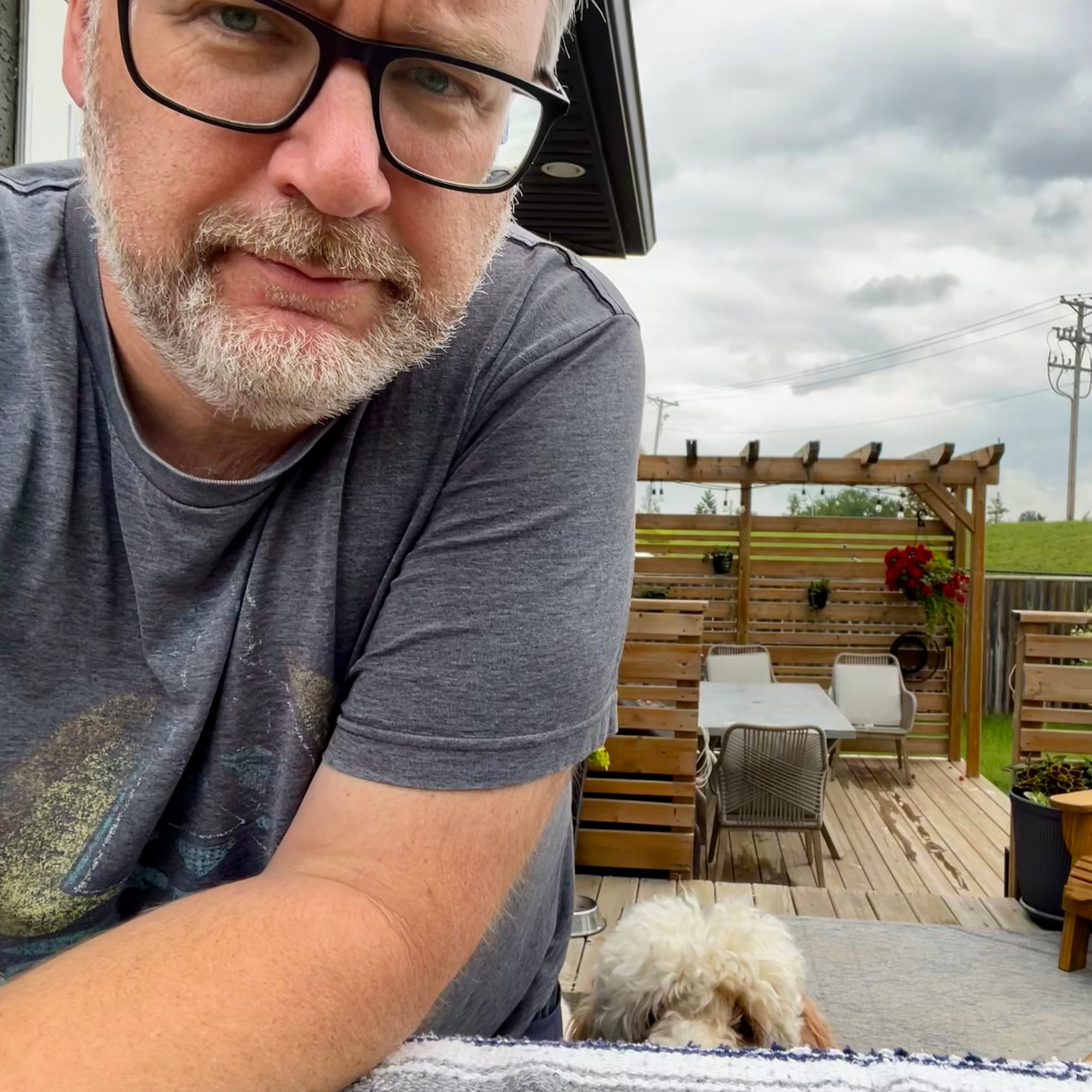 A man wearing glasses and a gray t-shirt is leaning forward while a small white dog peeks over a surface in an outdoor patio area with wooden furniture and plants.