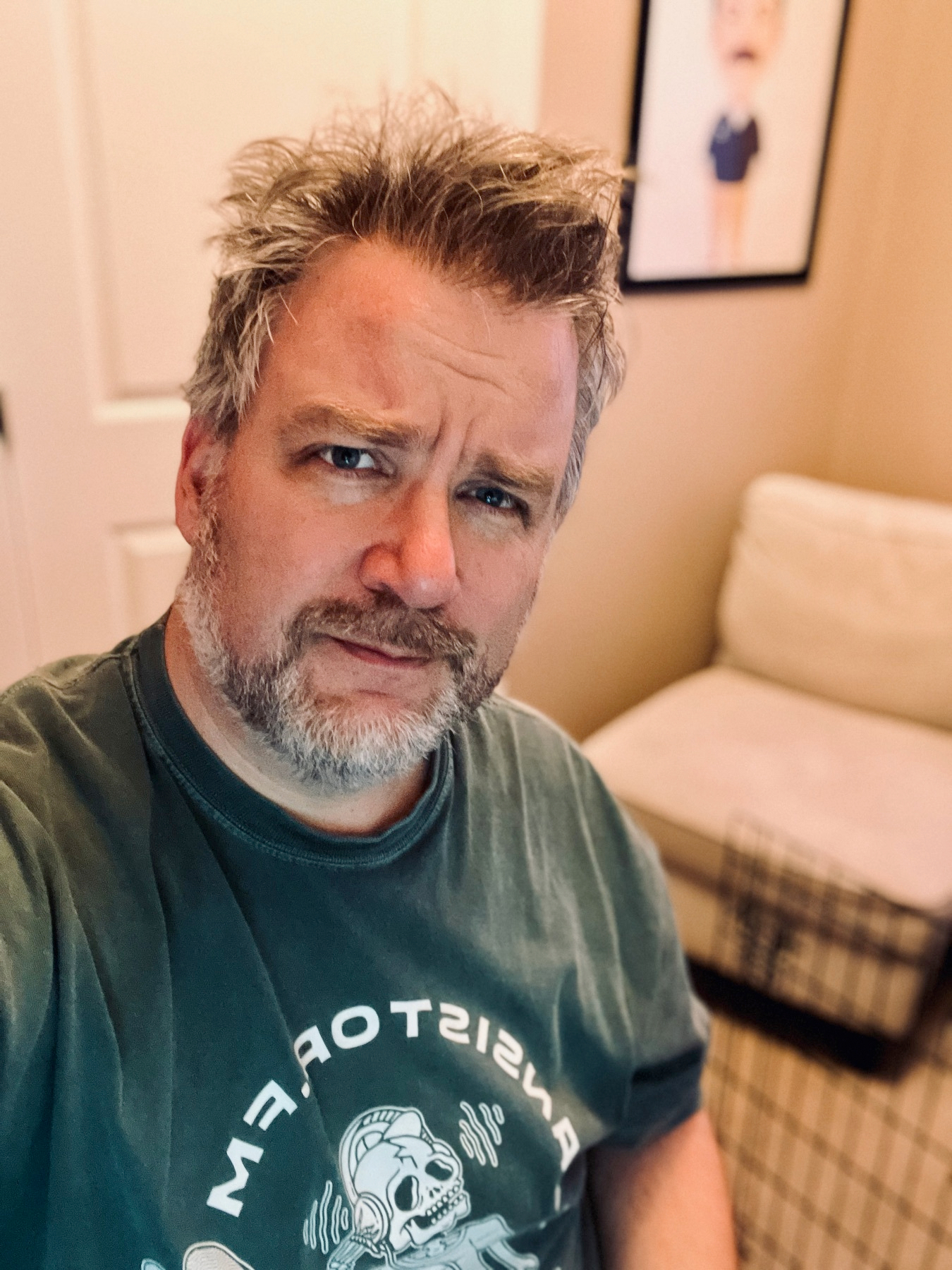 A man with tousled hair and a beard is sitting in a room with a light-colored chair and a framed picture on the wall.