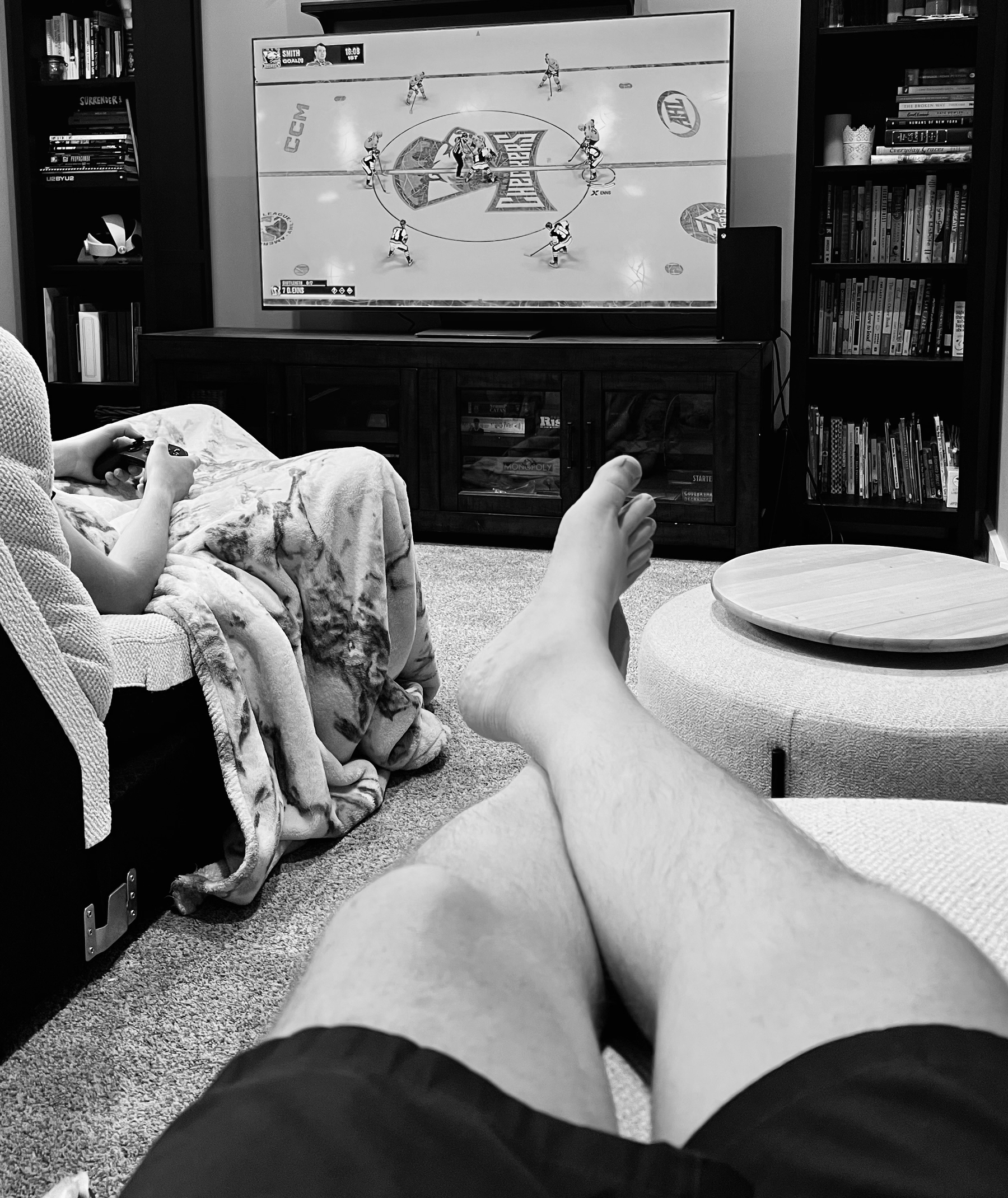 Two people are relaxing in a living room watching a hockey game on TV, with one person holding a gaming controller and the other person's legs visible in the foreground.