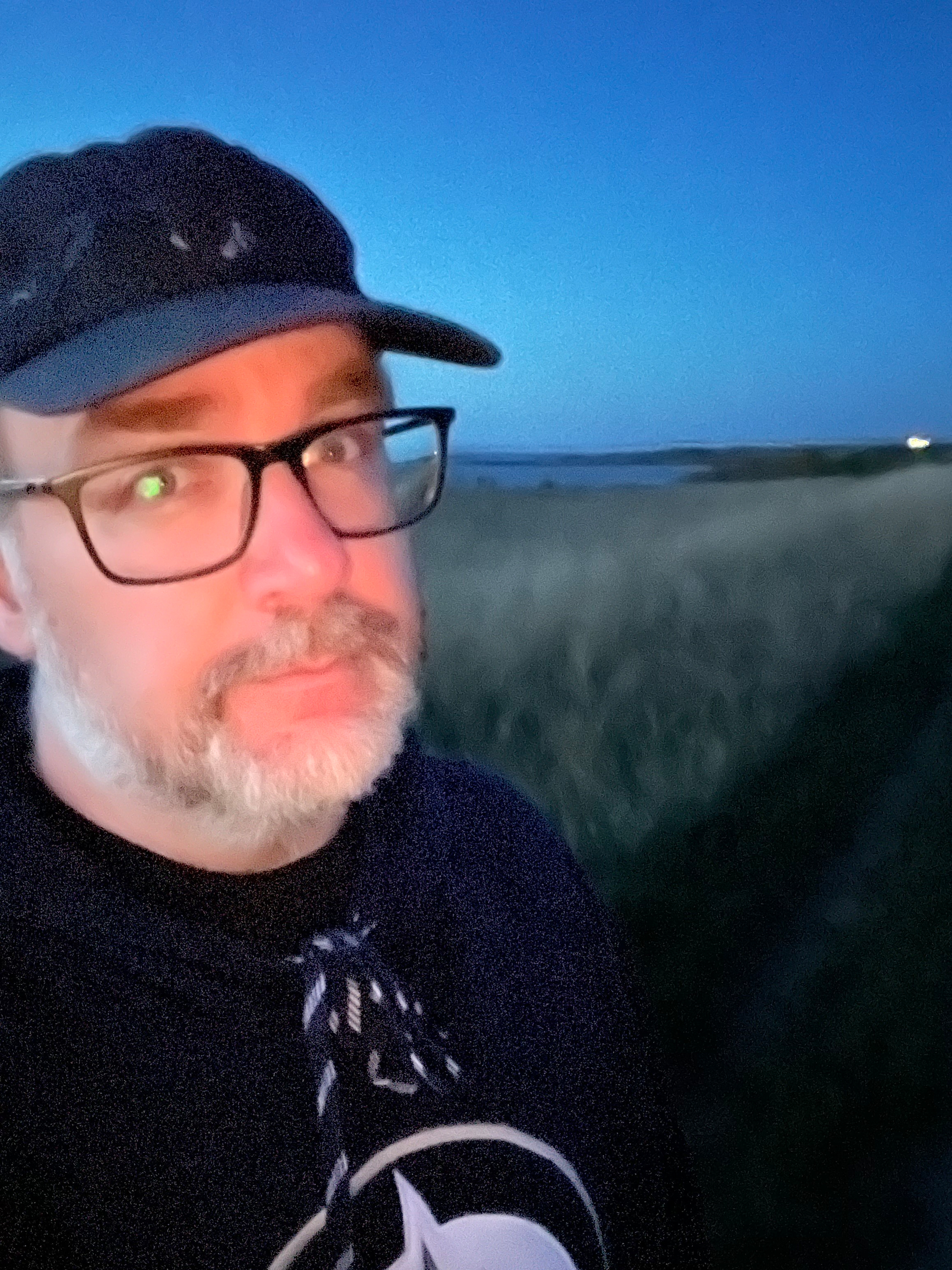 A person wearing glasses and a cap is taking a selfie outdoors at dusk, with a grassy field in the background.