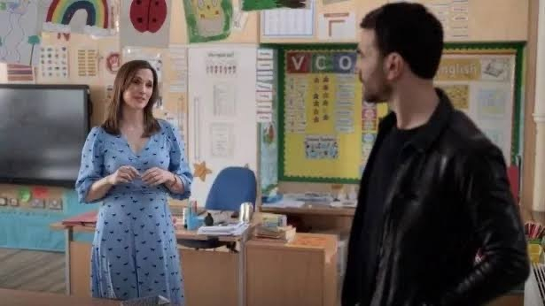 Ruth Bradley in a blue dress and Roy Kent in a black jacket are engaged in conversation inside a colorful classroom.