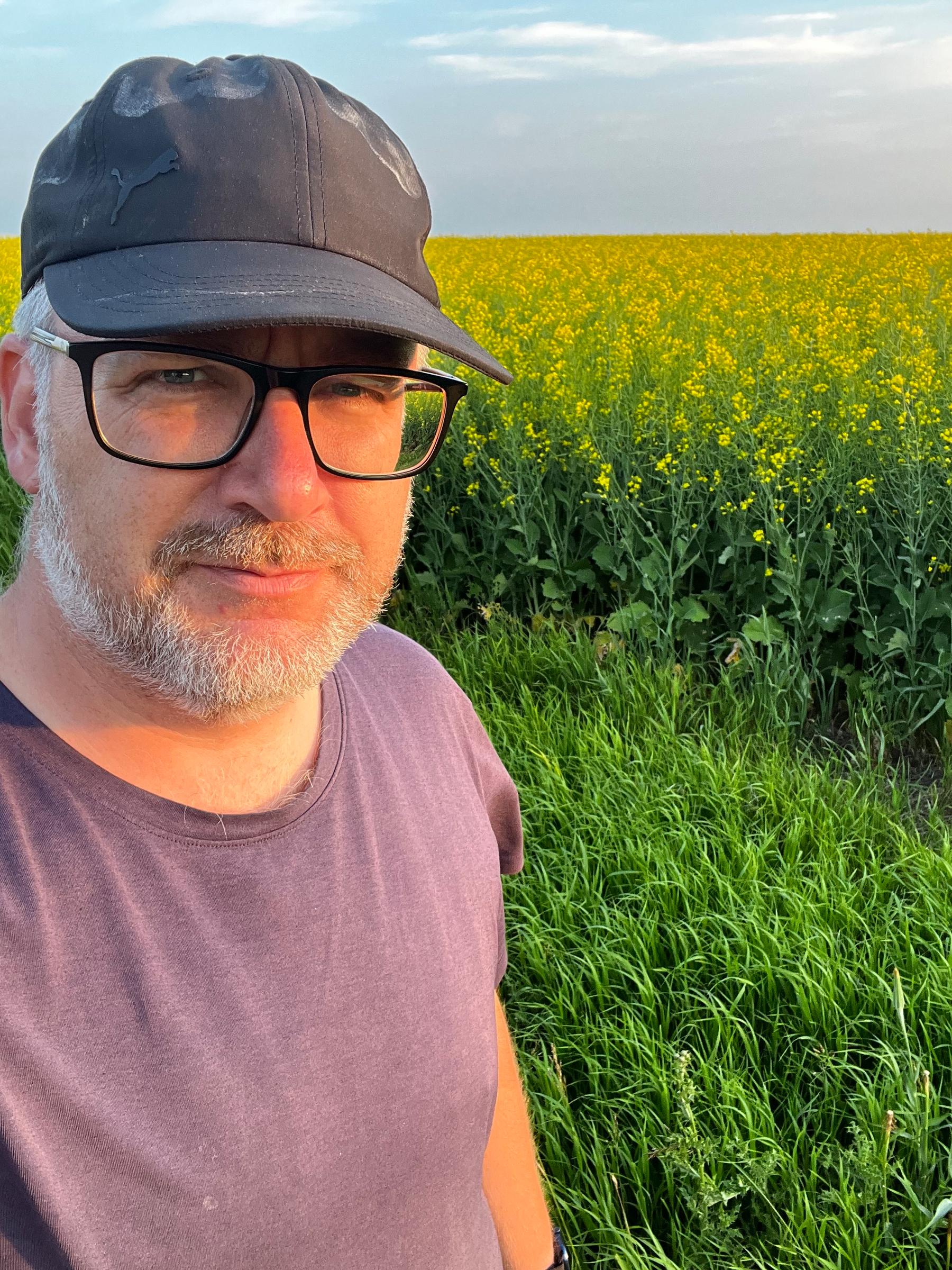 A man wearing glasses, a black cap, and a purple shirt is standing in front of a field with yellow flowers and green grass under a partly cloudy sky.