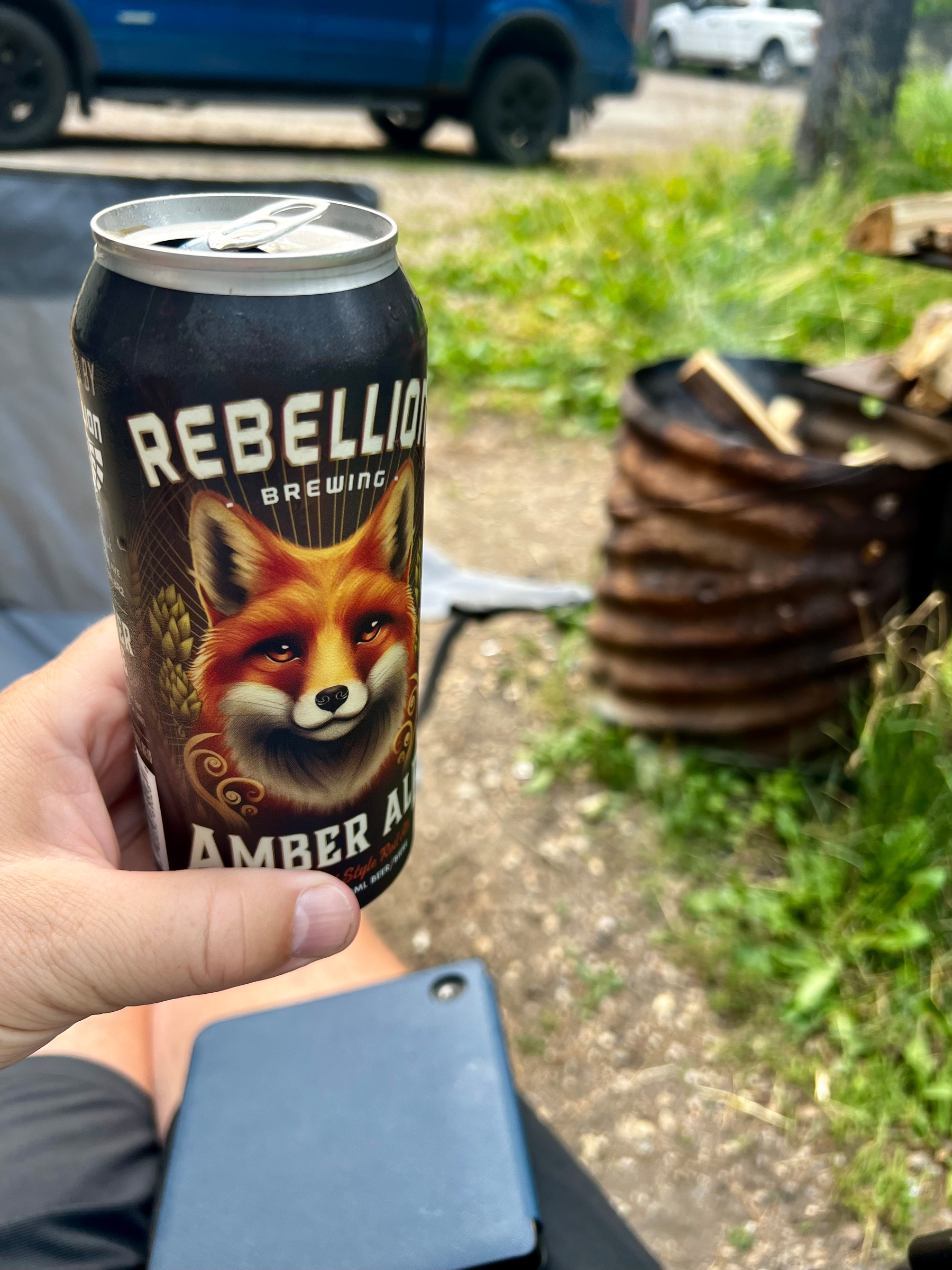 A person is holding a can of Rebellion Brewing Amber Ale near a campsite with a fire pit and some greenery.