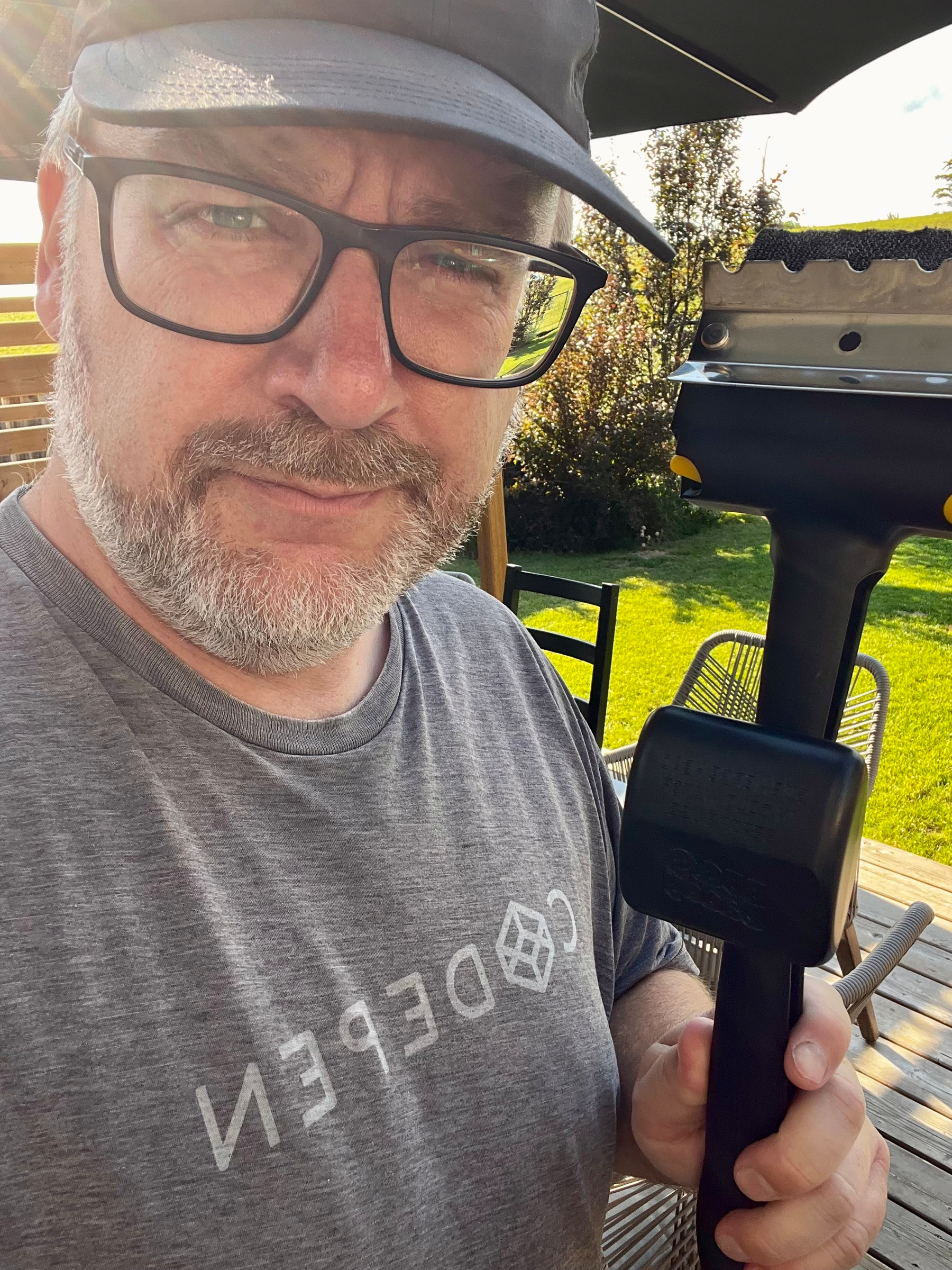 A man wearing glasses and a cap is holding a handheld grill scraper while standing on a wooden deck with outdoor furniture and greenery in the background.