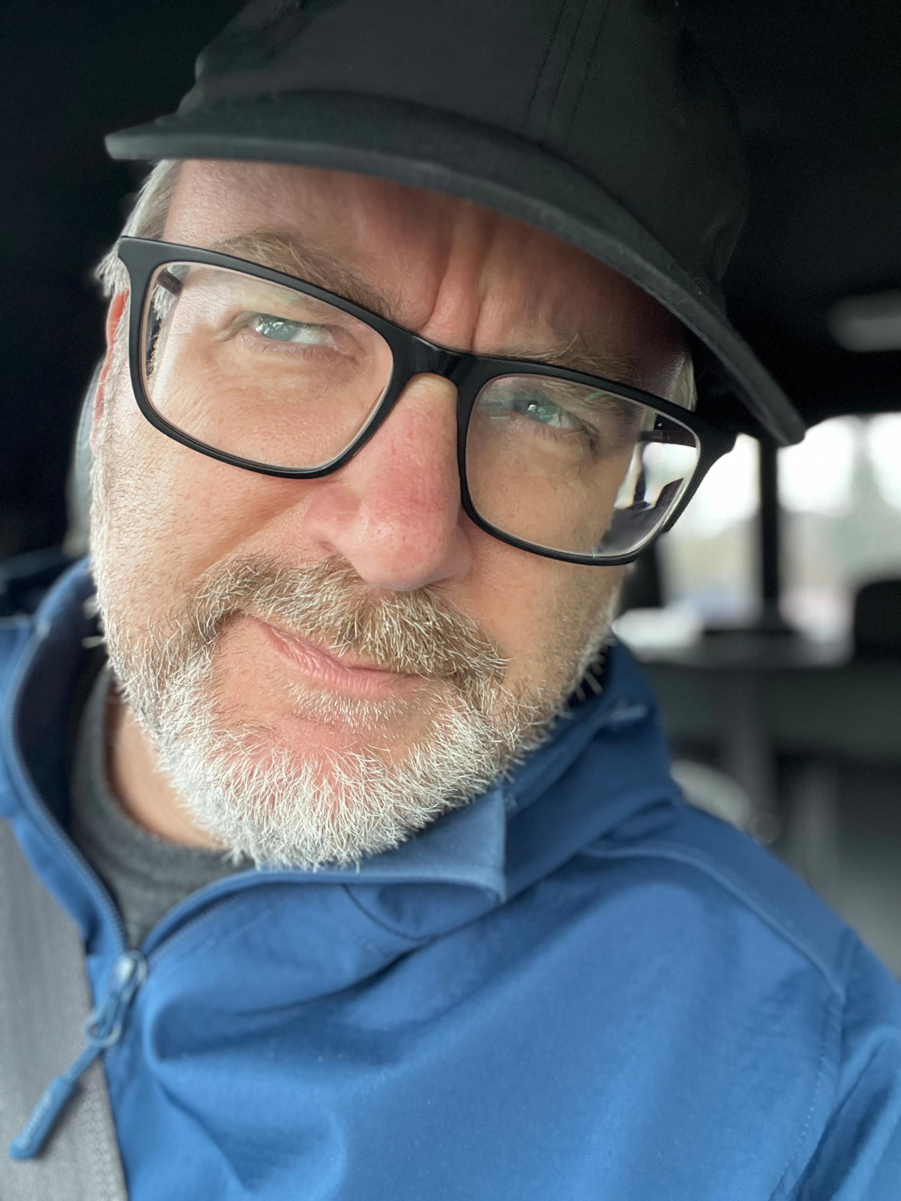 A man with glasses and a cap is sitting in a vehicle, wearing a blue jacket and looking at the camera.