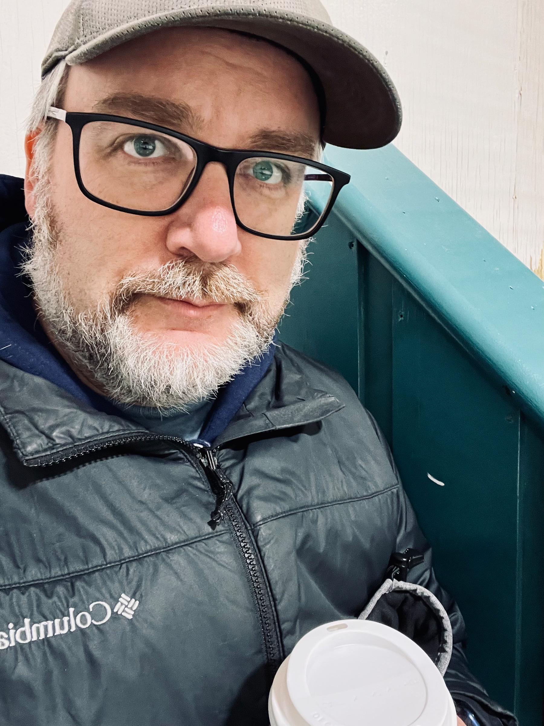A man with glasses and a cap is holding a coffee cup while wearing a dark jacket.