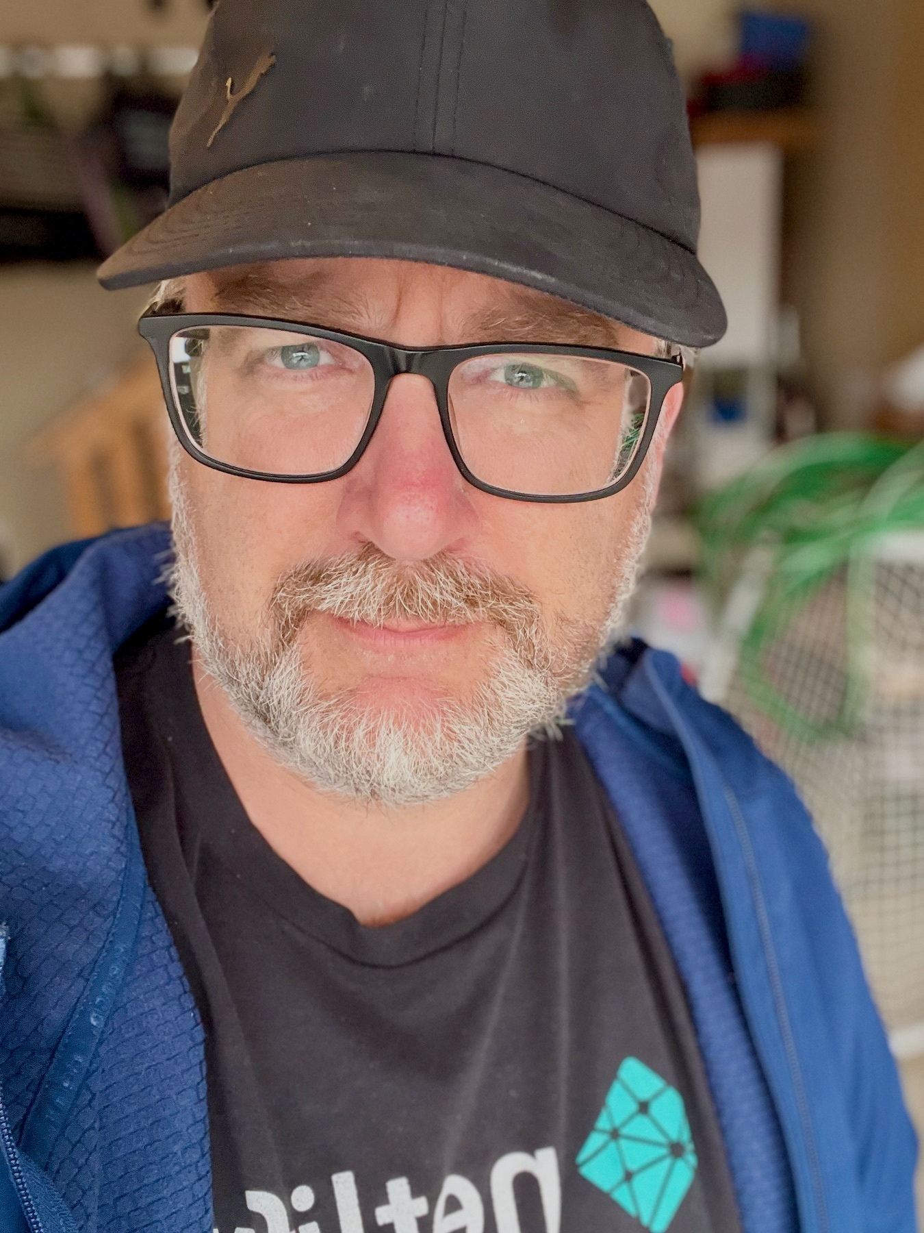 A bearded man wearing glasses, a black cap, and a blue jacket stands in a room with various blurred background items.