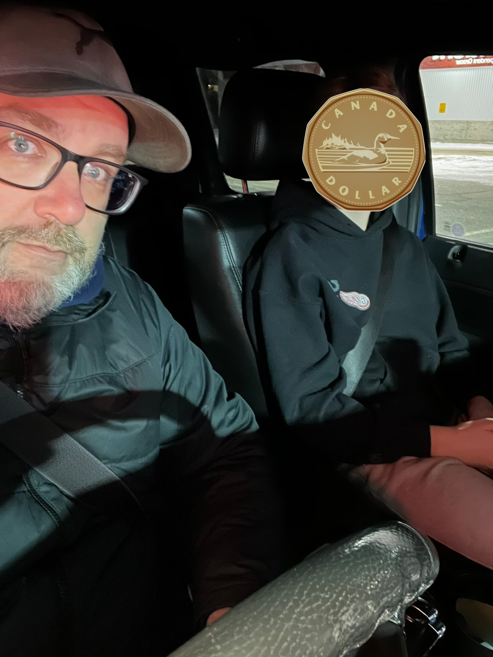 Two people are sitting inside a vehicle, with one person's face obscured by an image of a Canadian dollar coin.
