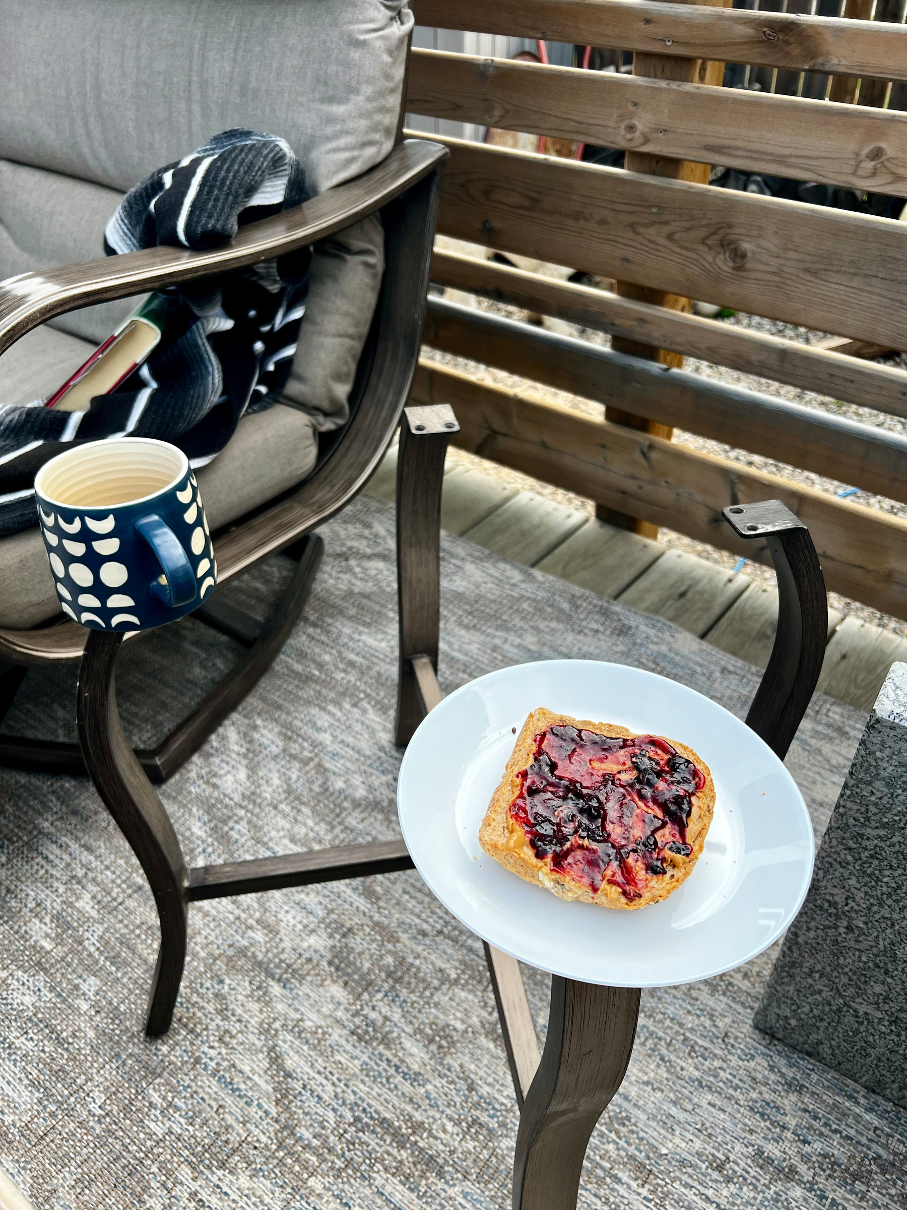 A cozy outdoor seating area features a cushioned wooden chair with a striped blanket, a patterned mug resting on the armrest, and a white plate holding a piece of toast with berry jam atop a metal side table.
