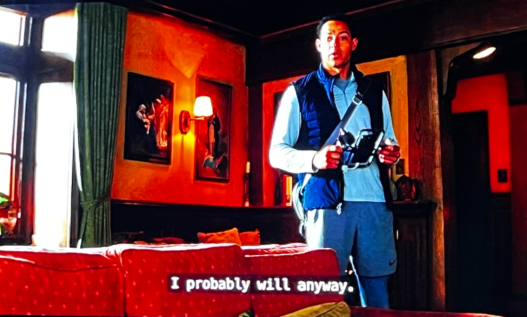 A person stands in a warmly decorated room with art on the walls, wearing casual clothing and holding a device. Subtitle says I probably will anyway. 