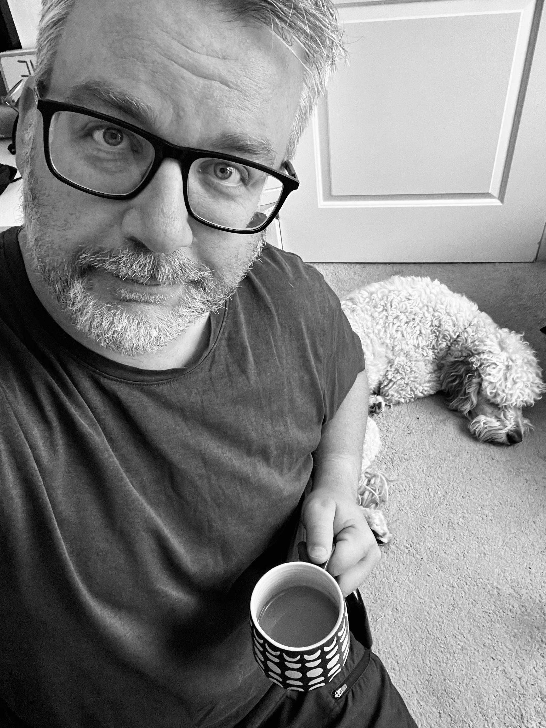 A person (me!) is holding a mug of coffee is sitting next to a sleeping dog on the floor.