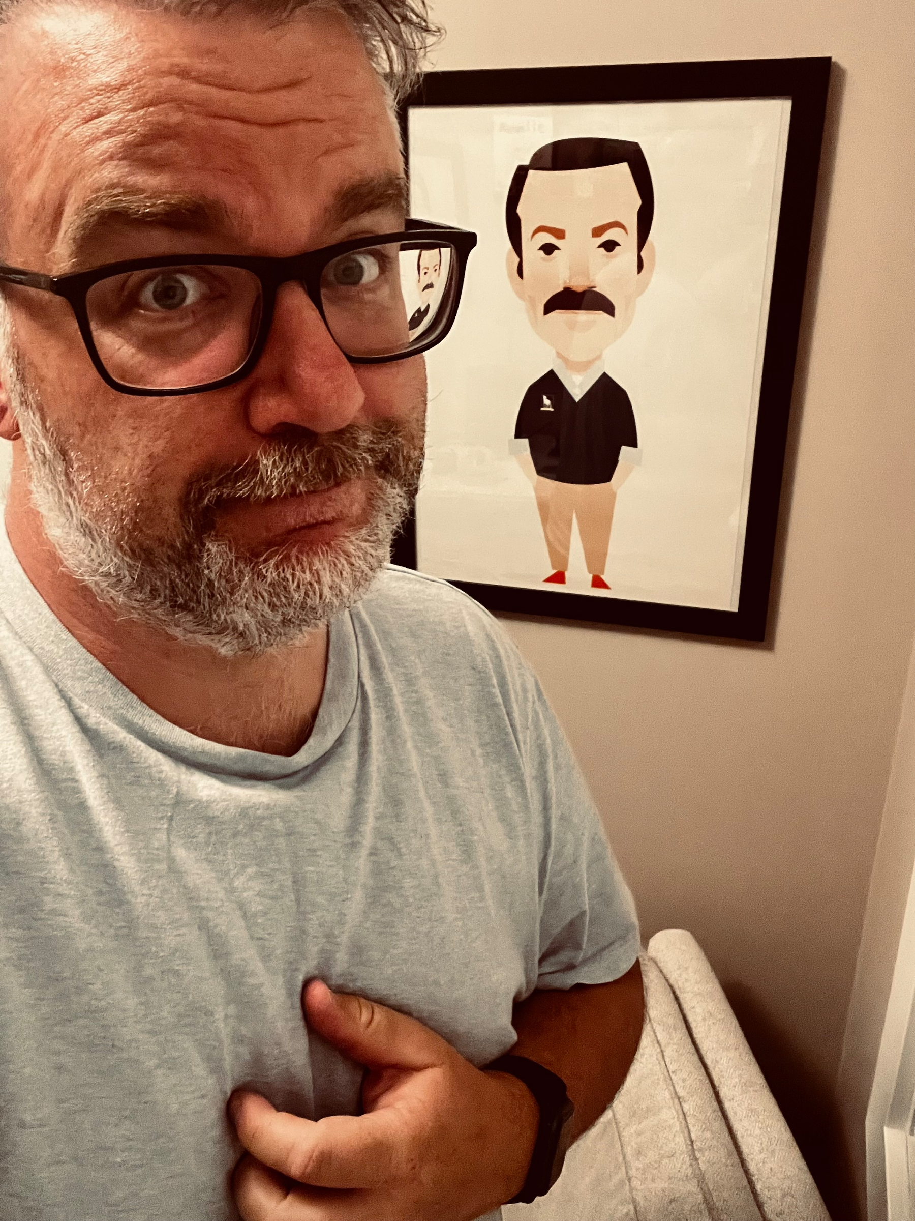 A bearded man wearing glasses and a light blue shirt poses with a slight smile in front of a caricature of Ted Lasso framed on the wall.