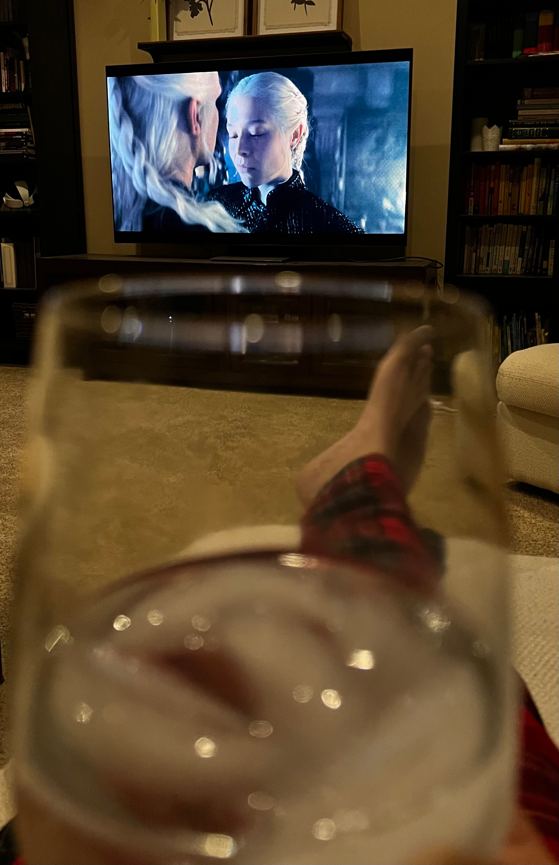 A person wearing red plaid pants is relaxing with a drink in hand while watching a scene from a television show featuring two characters with white hair.