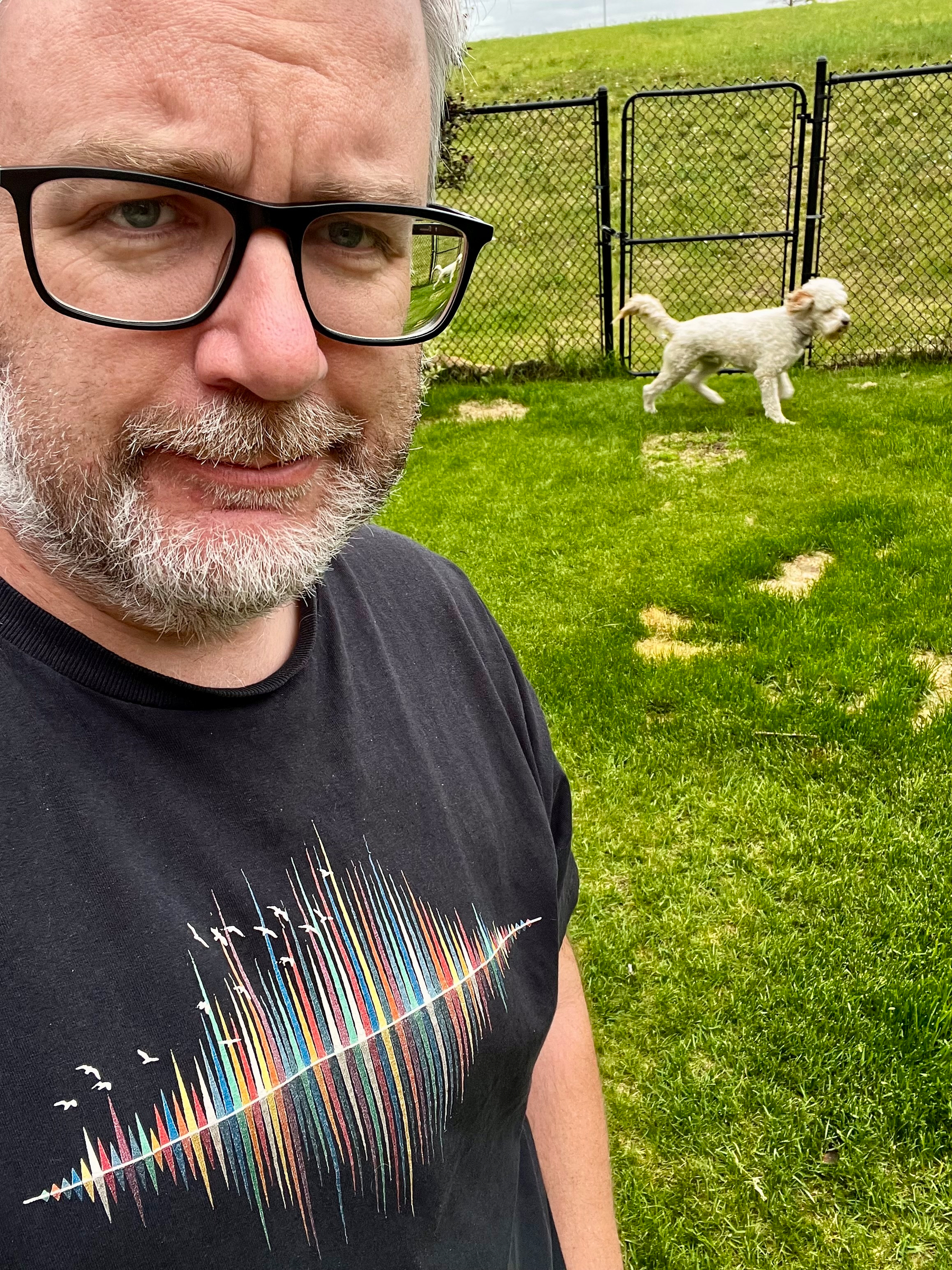 A man with glasses and a T-shirt featuring colorful lines stands on grass, while a white dog walks near a fence in the background.
