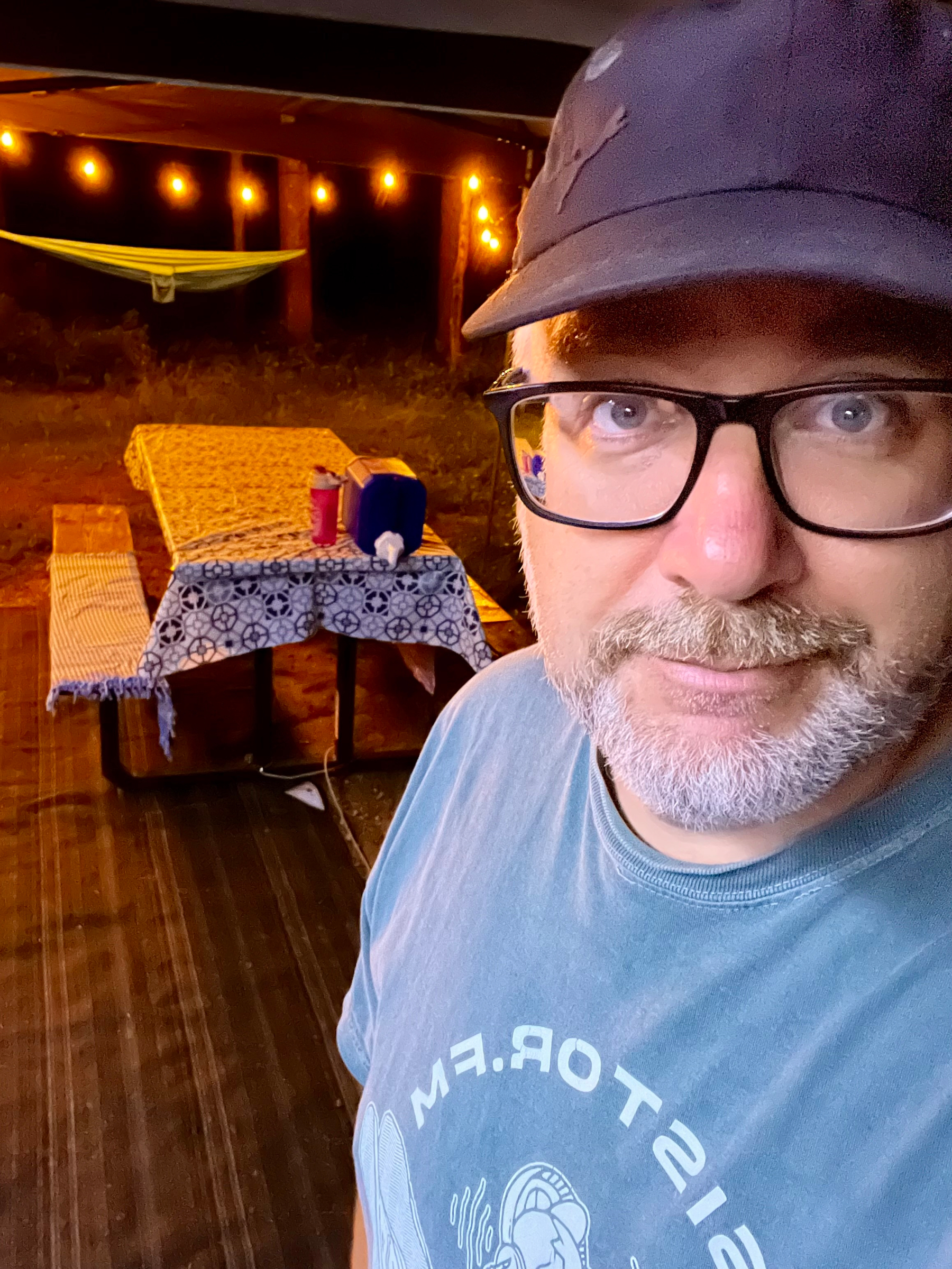 A man wearing glasses and a cap takes a selfie near a picnic table covered with patterned cloth and illuminated by string lights in an outdoor setting at night.